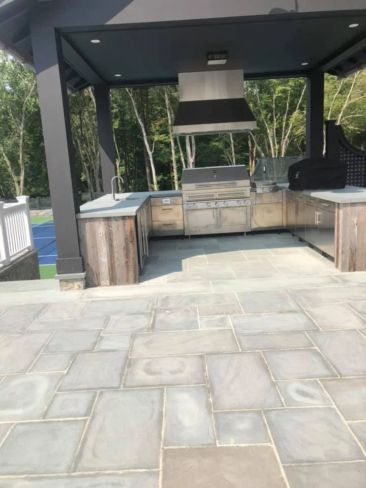 Outdoor kitchen with a grill, a hood, and cabinetry, under a covered patio with stone flooring and surrounded by trees.