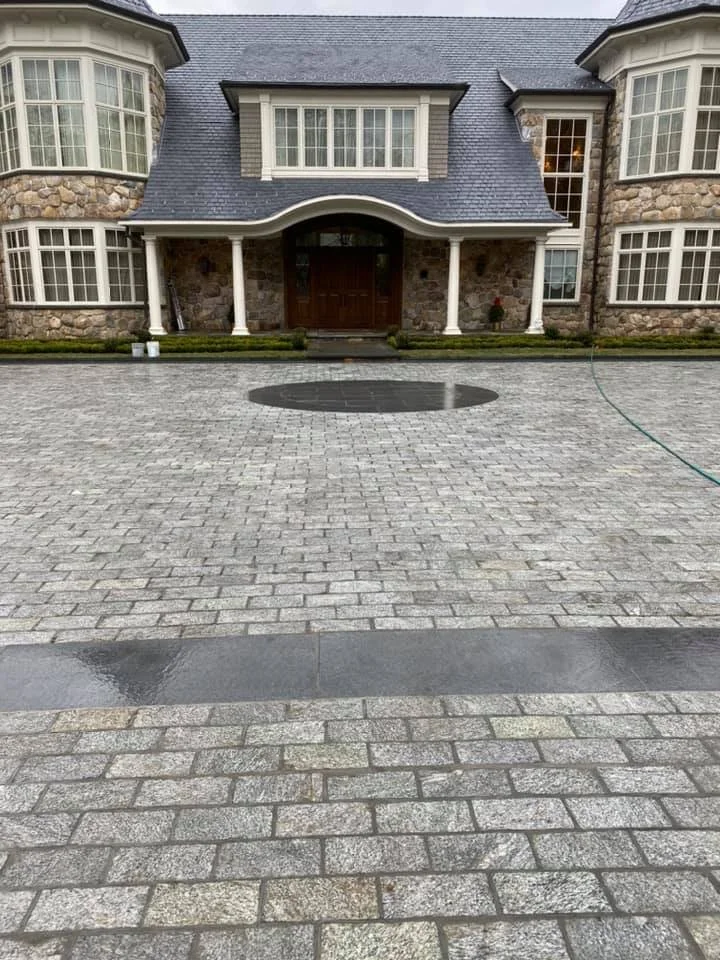 A large house with stone and siding exterior, multiple windows, and a front porch with columns. The driveway in front is paved with light-colored bricks and has sections of black stone. A garden hose is on the right side of the driveway, and there ar