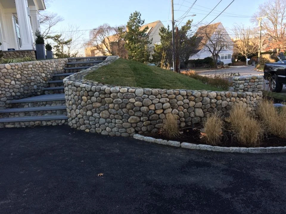 Stone retaining wall with steps leading up to a grassy yard, surrounded by a landscaped area with ornamental grasses, in a suburban neighborhood with houses, trees, and utility poles.