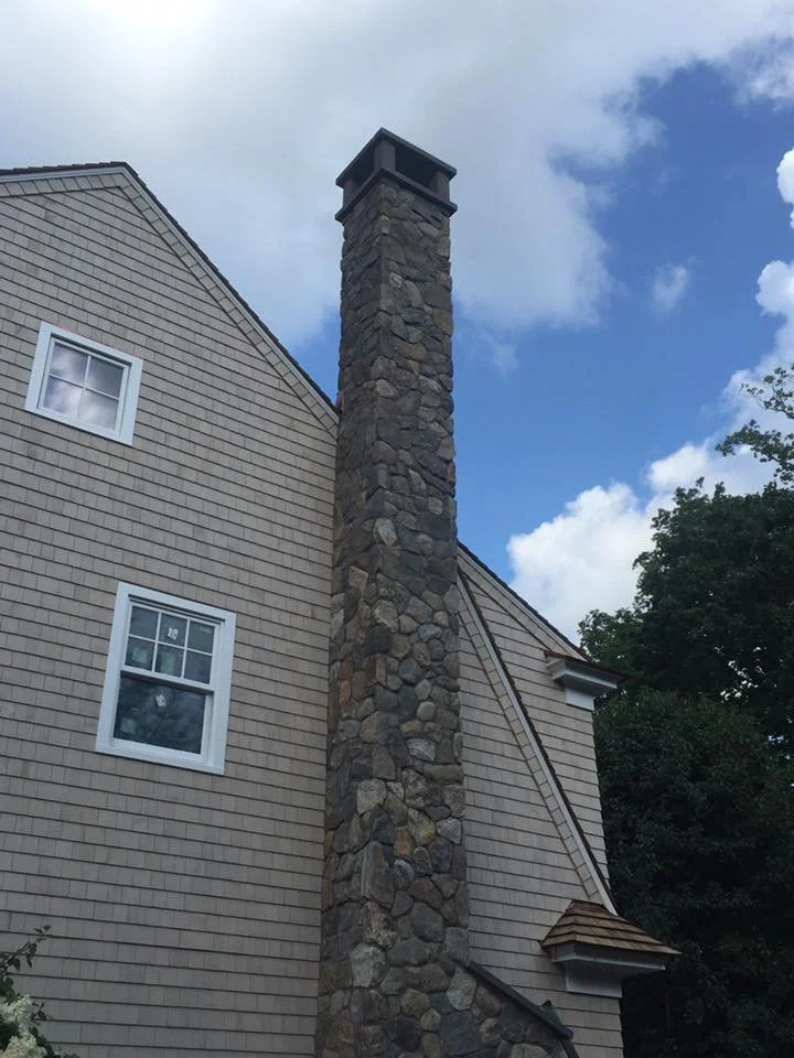 View of a house exterior with a stone chimney and beige siding under a partly cloudy sky.