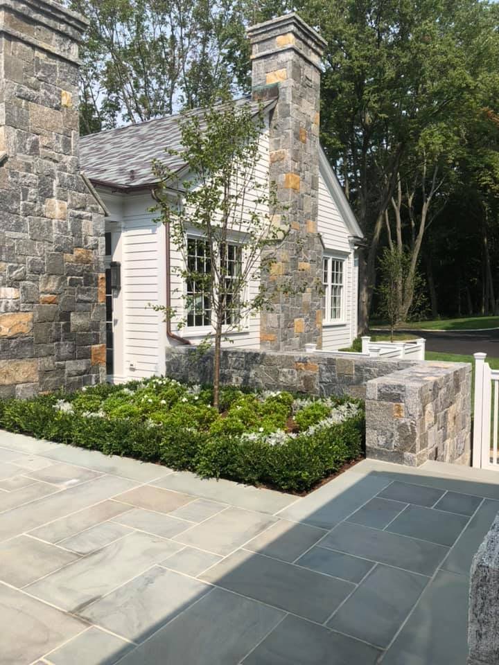 A house exterior with stone and white siding, large windows, a small tree, and a landscaped garden with shrubs and flowers. Stone pathway in the foreground.