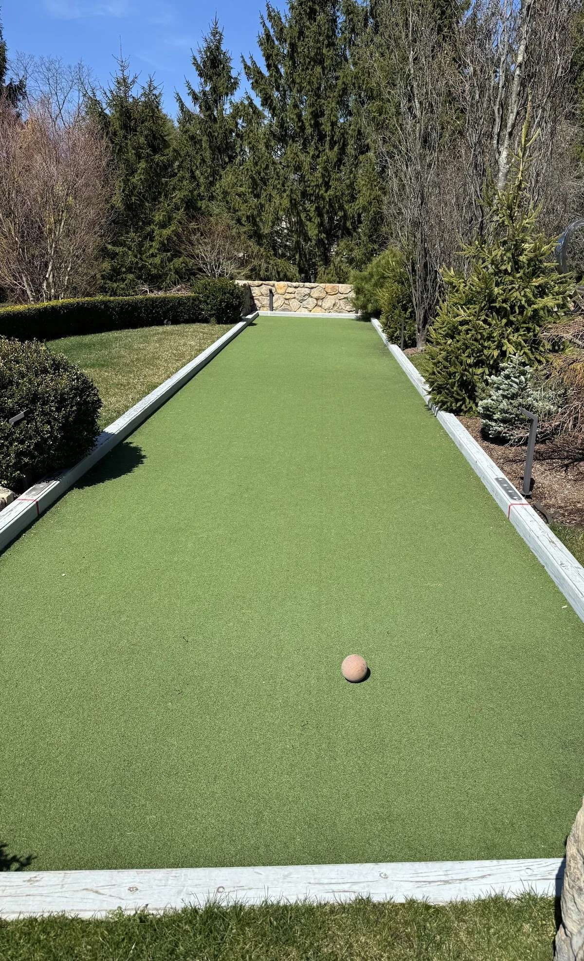 A backyard with a miniature golf putting green, small shrubs, trees, and a stone wall in the background.