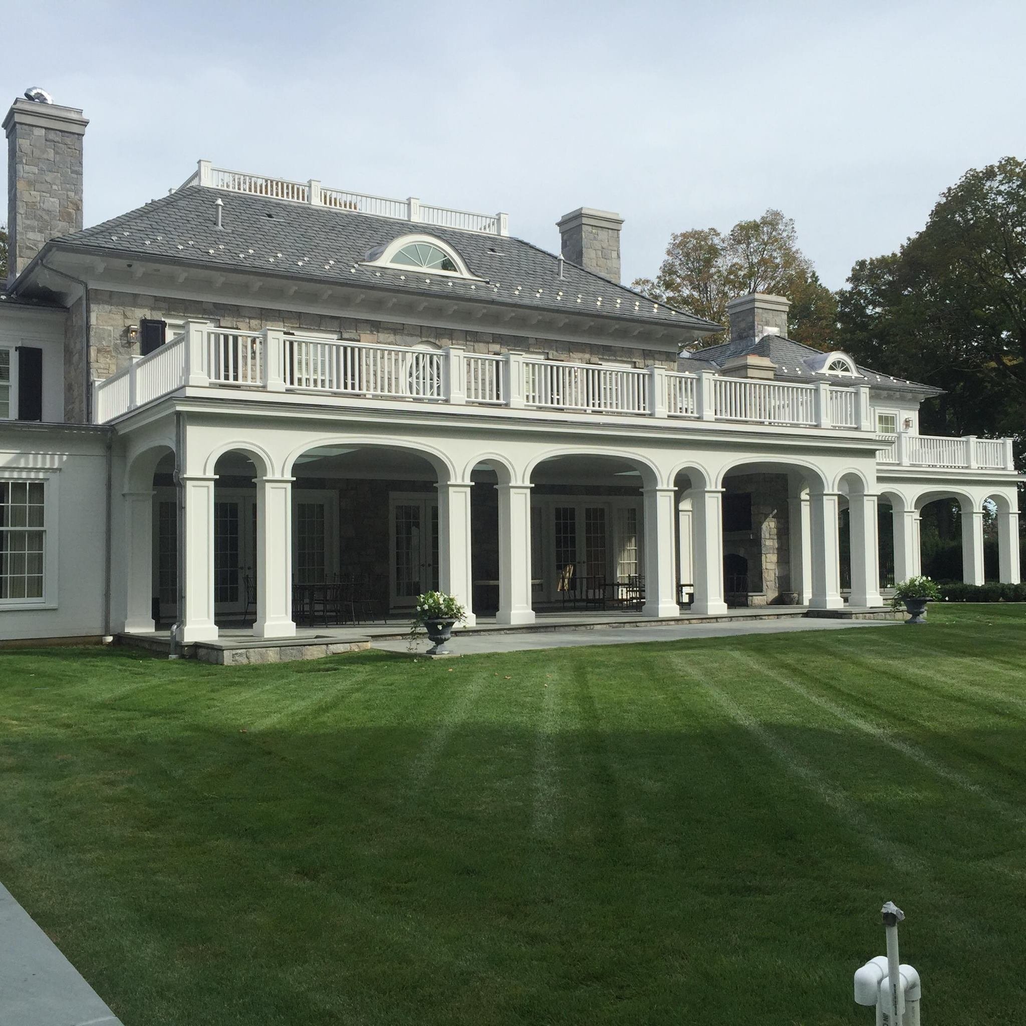 Large, elegant house with multiple levels, stone and white exterior, with arches on the ground level and balconies on the upper level, surrounded by a well-manicured lawn.