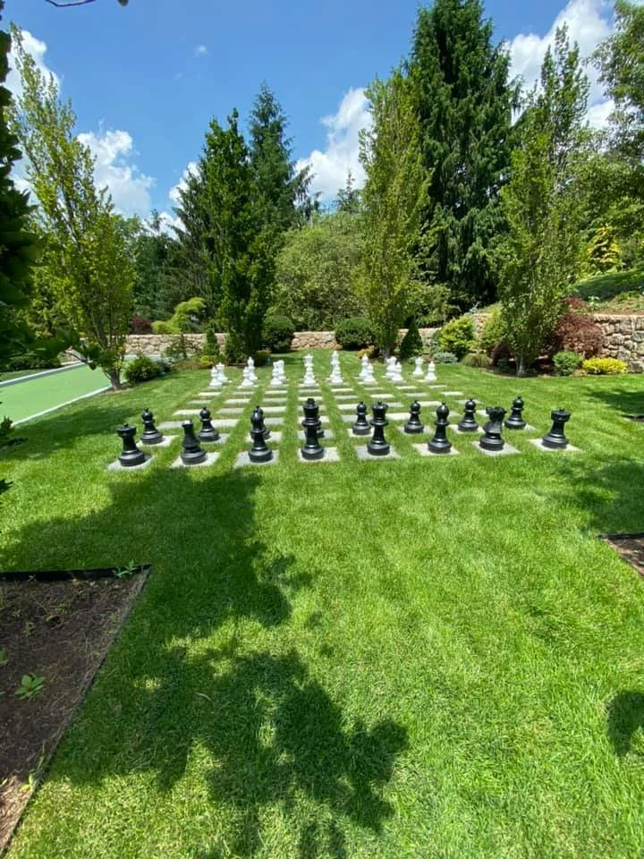 Large outdoor chess set with black and white pieces on a grass lawn, surrounded by trees and a stone wall, under a partly cloudy sky.