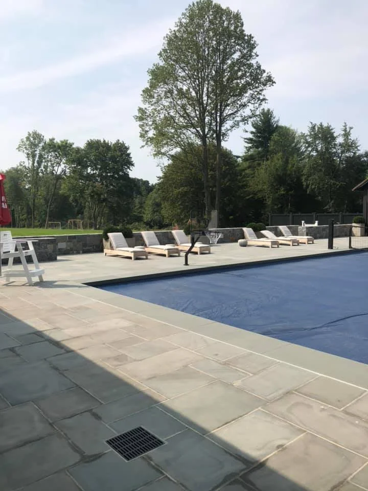Empty swimming pool covered with a dark blue cover, with poolside lounge chairs and a basketball hoop in the background, surrounded by trees and a cloudy sky.