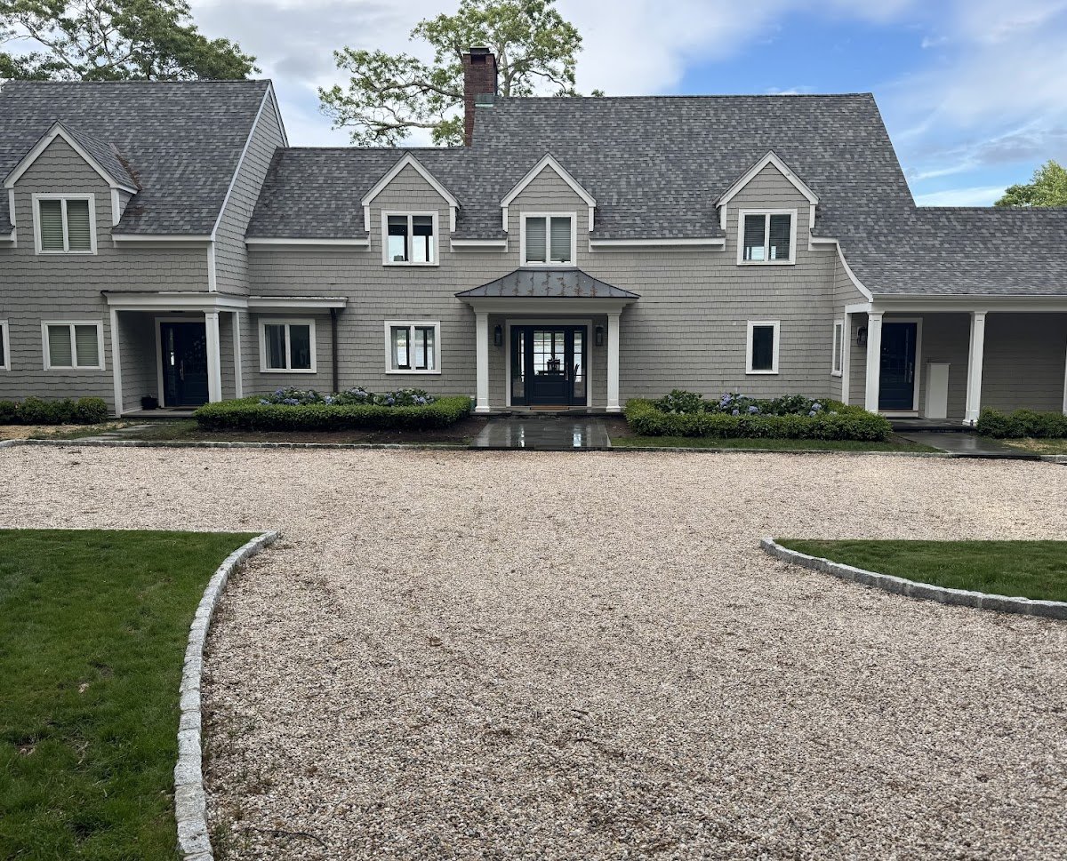 A large gray house with multiple windows, a steep roof, and a chimney. It has a front porch with columns and a gravel driveway leading to the entrance, flanked by green lawns and shrubbery.