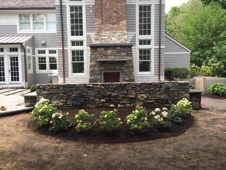A stone and brick outdoor fireplace with a landscaped flower bed in front of it, adjacent to a residential house with grey siding and white trim, and a backyard with trees and a wooden fence.