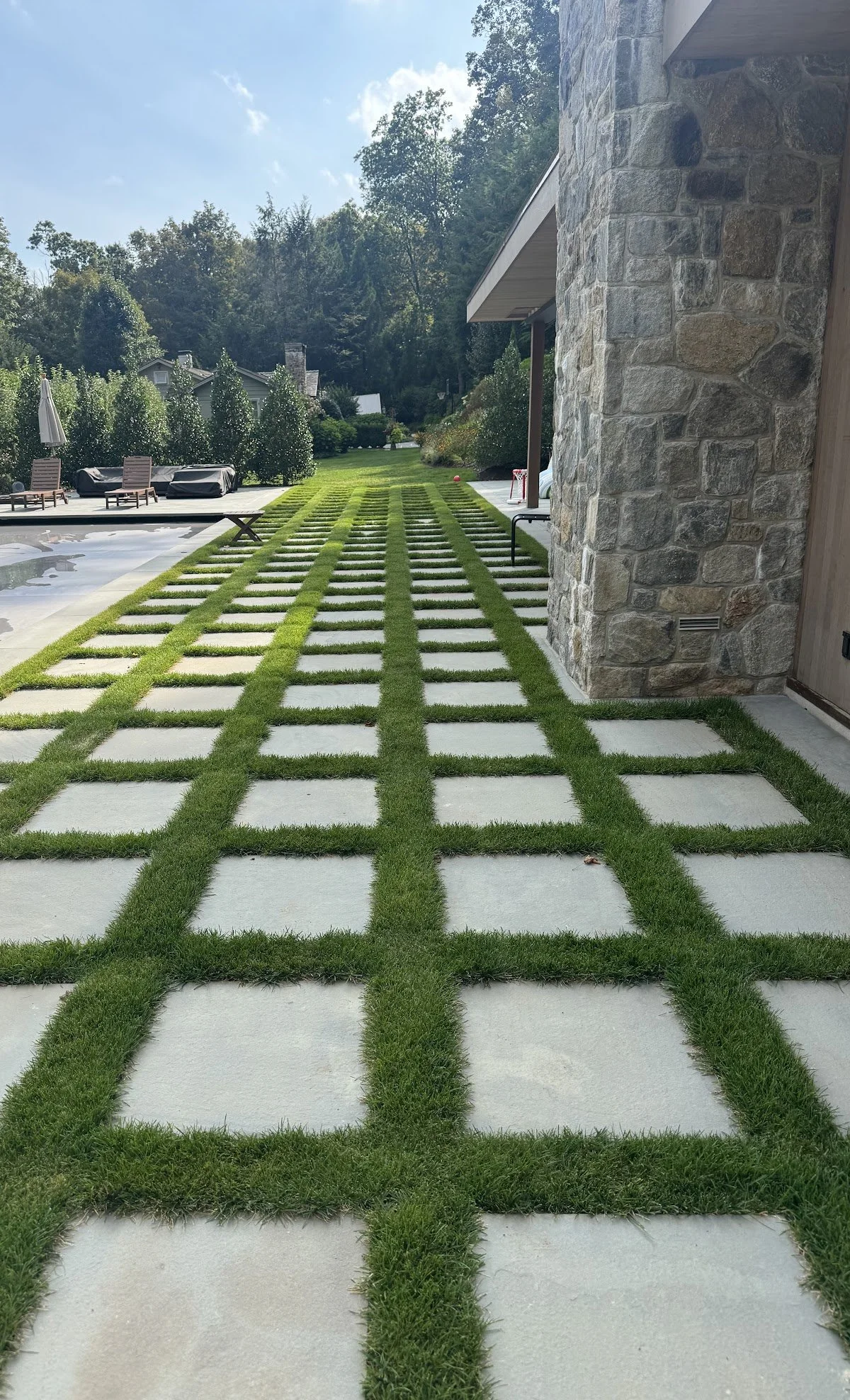Modern house exterior with a stone wall, a checkered pattern of concrete squares and grass on the walkway, surrounded by green lawn and trees.
