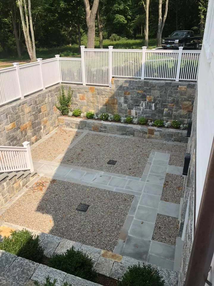A backyard with a stone and gravel patio, enclosed by white fence, with mature trees in the background and a black vehicle parked nearby.