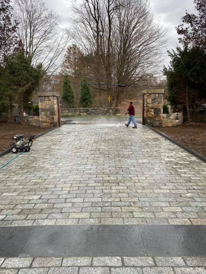 A person pressure washing a brick driveway near stone gate pillars and trees in the background.