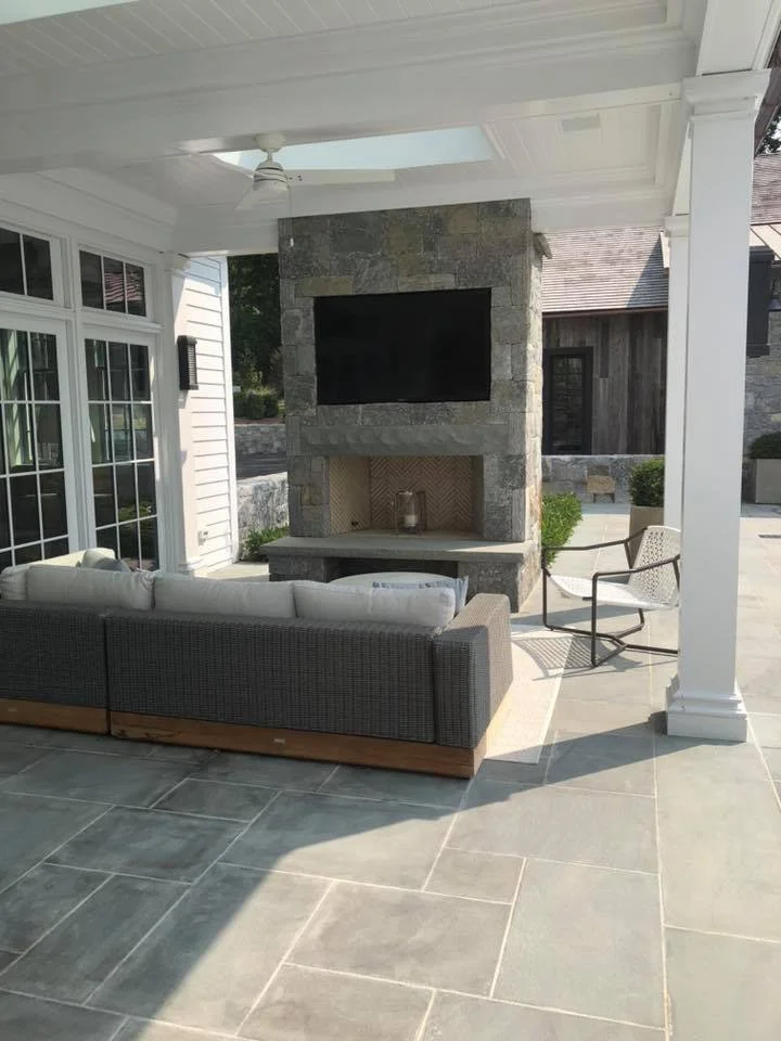 Outdoor patio with a gray sectional sofa, a white metal chair, a stone fireplace, and a large TV mounted above the fireplace, under a white ceiling with a ceiling fan.