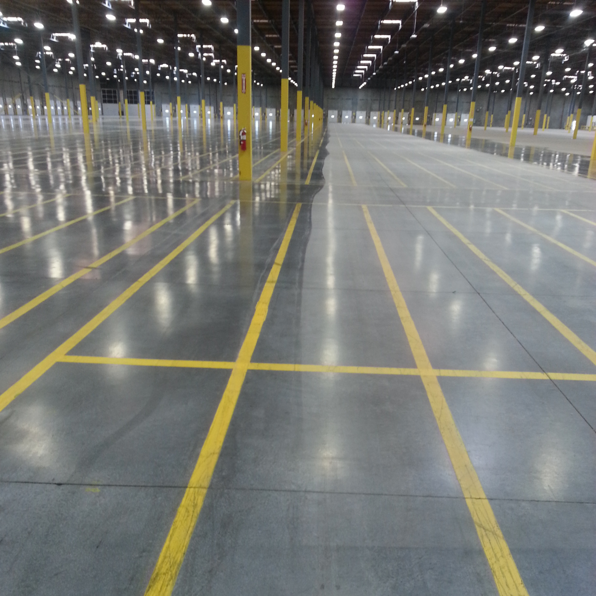 Empty warehouse with yellow lines on the gray concrete floor and yellow safety poles dividing the space. Overhead lights illuminate the high ceiling and the walls have large garage doors.
