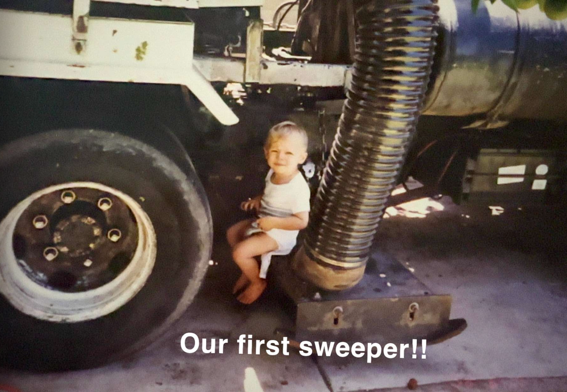 A young child sitting on a wooden platform next to a large industrial vacuum cleaner with a corrugated hose, smiling at the camera with the words "Our first sweeper!!" overlaid on the photo.