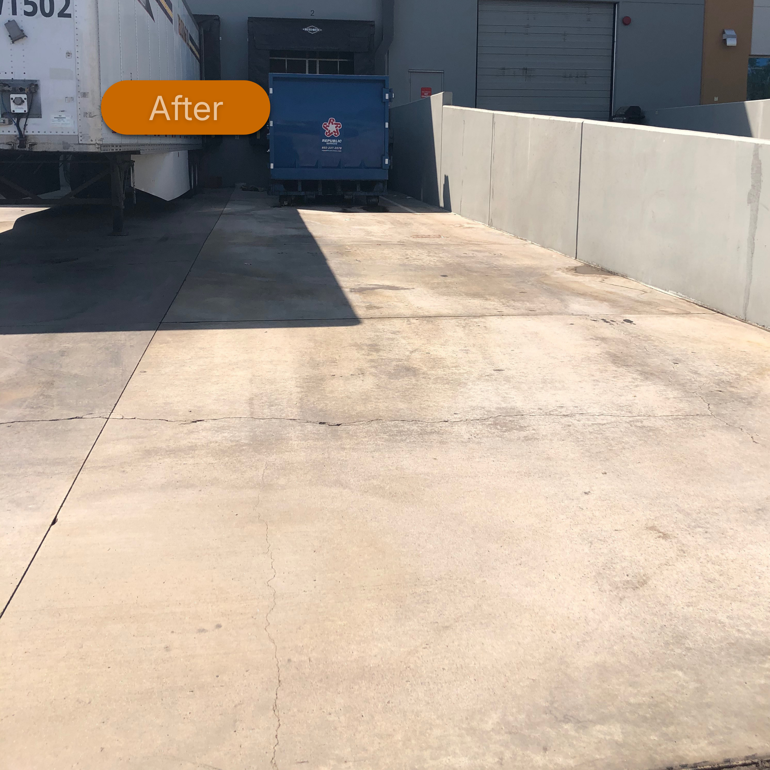 A concrete loading dock area with a large white truck on the left and a blue trash bin in the middle near a beige building. The photo is taken during the daytime with shadows cast on the ground.
