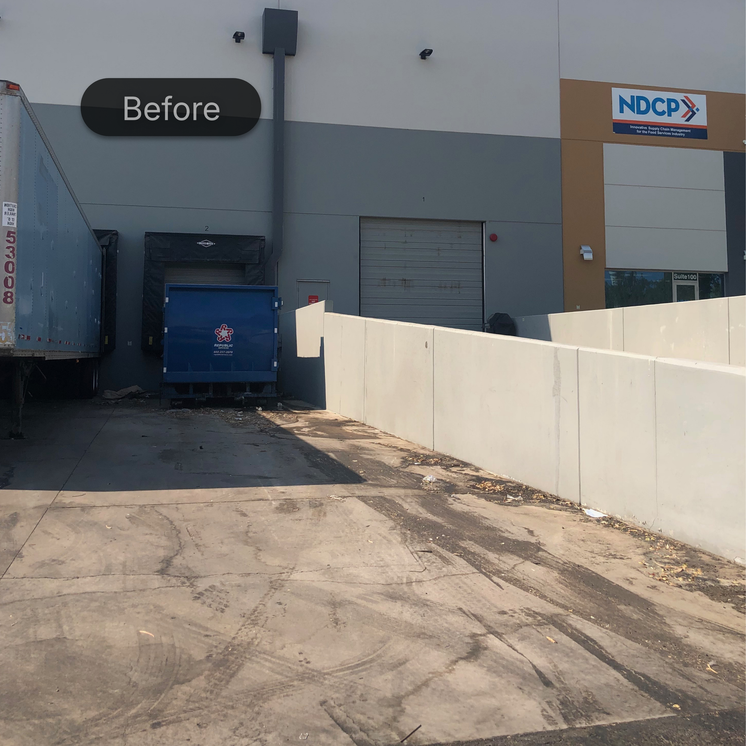 The image shows the exterior of a commercial building with a sign that reads NDCP and a slogan underneath. There are two large garage doors, a blue recycling bin, and a black trash bag visible in the area. The ground has some dirt and debris, and there is a white boundary wall in the foreground.