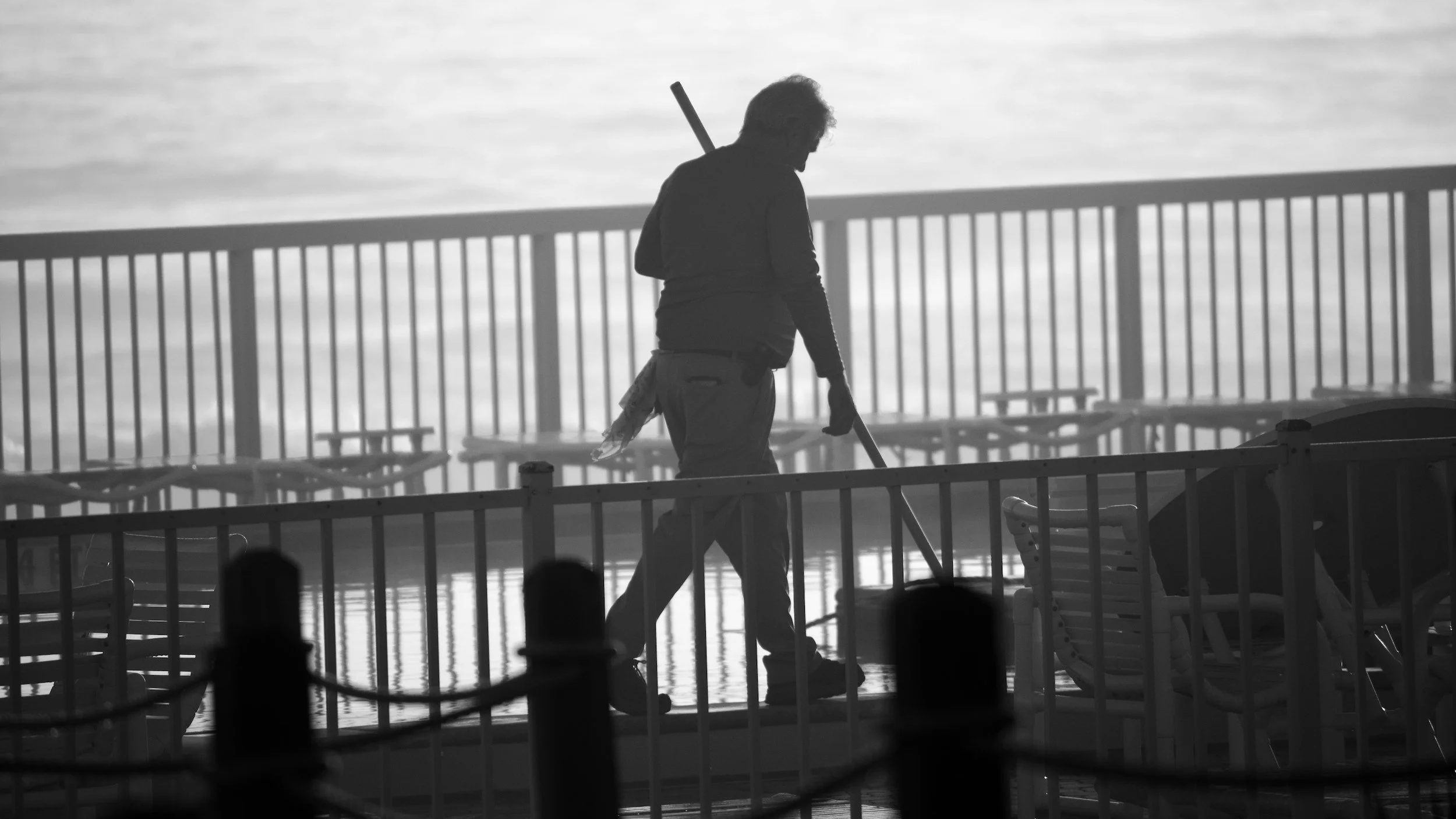 Silhouette of a person sweeping on a dock with empty chairs and a railing, water in the background, and a cloudy sky.