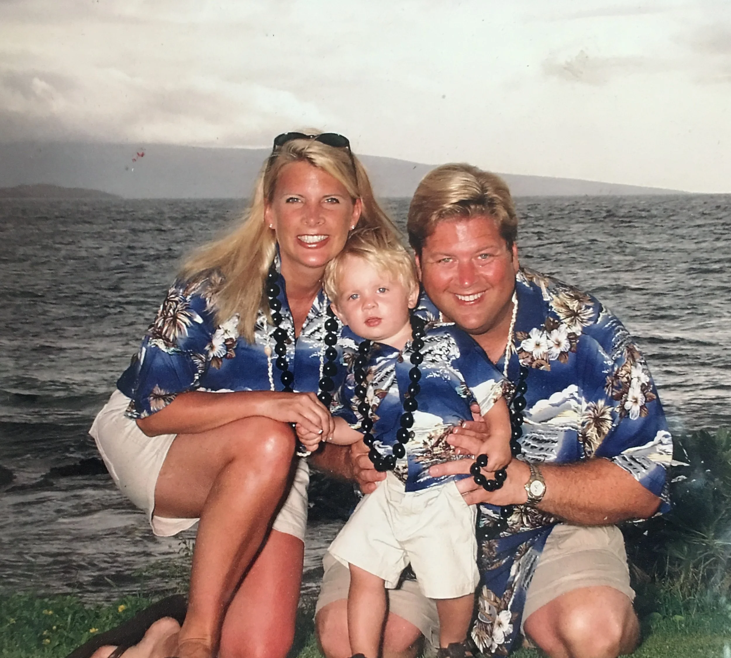 A family of three in Hawaiian shirts celebrating on a beach or lakeside with a large body of water in the background.