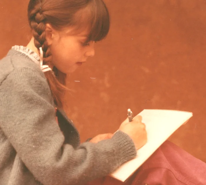 A young girl with braided hair, wearing a gray sweater, writing in a notebook with a pen against a reddish-brown background.