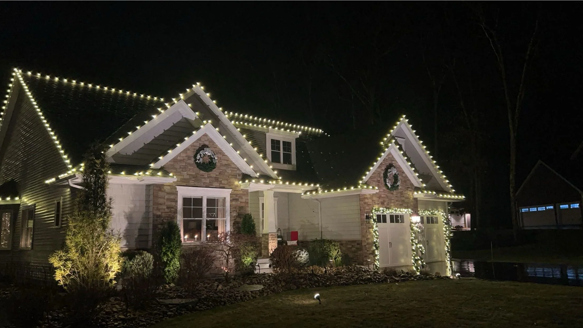 Nighttime photo of a house decorated with Christmas lights, including wreaths on the gables and illuminated garlands on the garage doors.