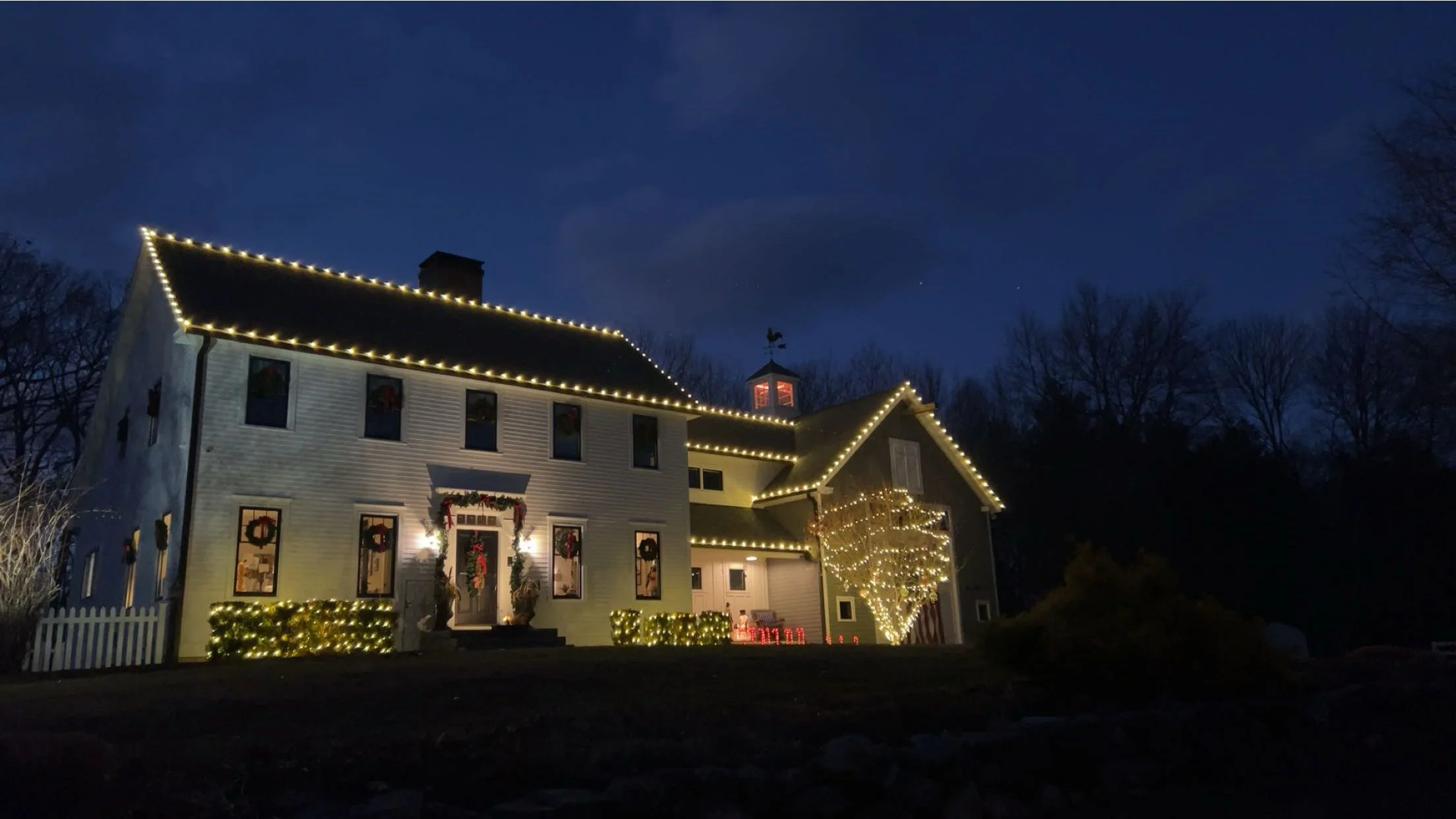 A two-story house decorated with Christmas lights during nighttime. The house has string lights outlining the roof and a leafless tree wrapped in lights in front. The windows are decorated with wreaths and the front yard has illuminated bushes.