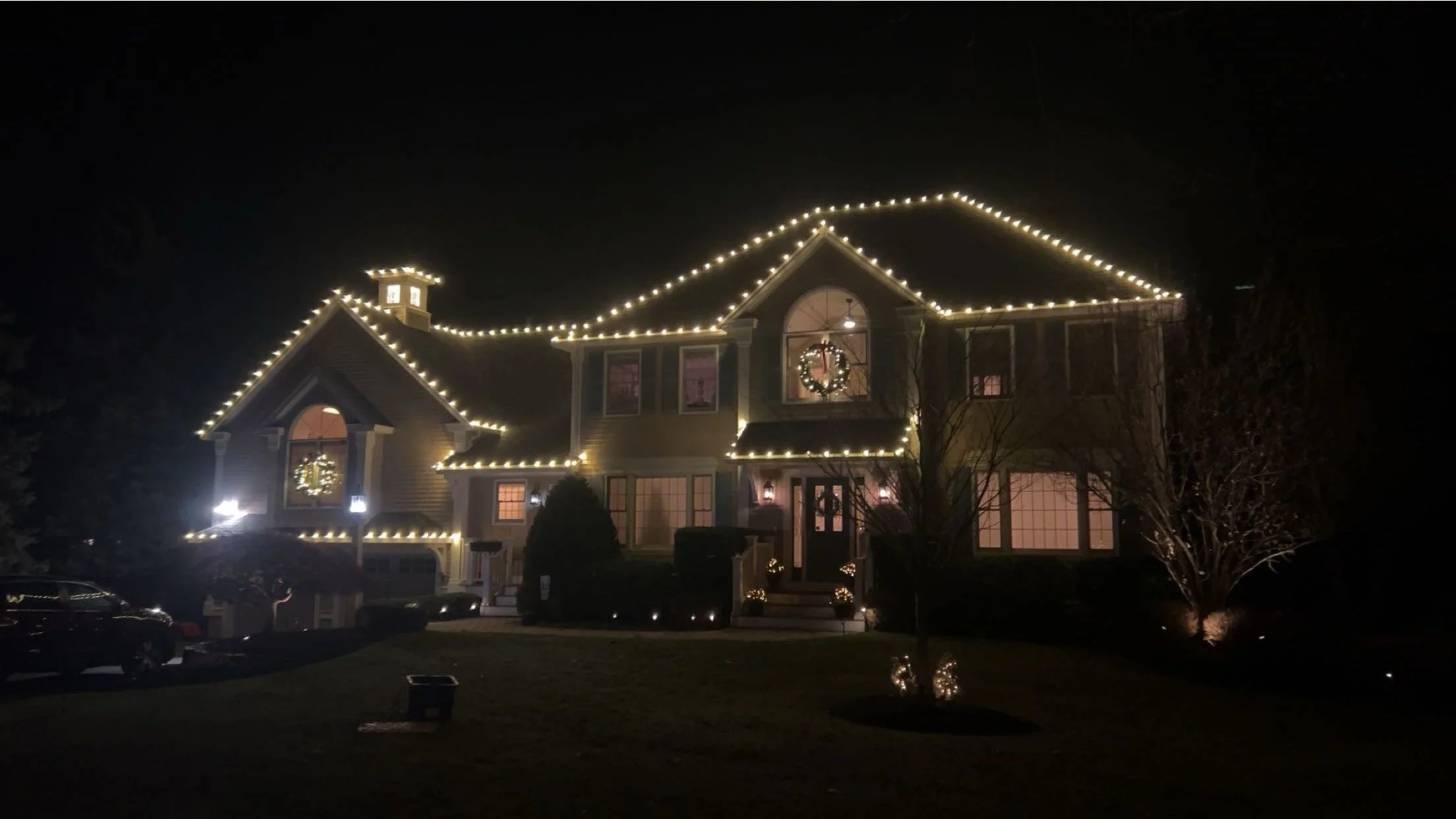 A house decorated with white Christmas lights outlining the roof and windows, with a festive wreath on the front door and illuminated decorations in the yard, during nighttime.