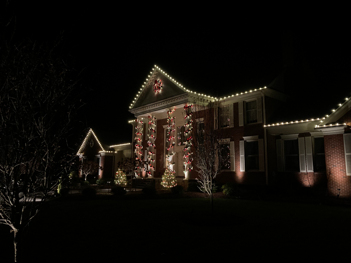 A house decorated with Christmas lights and ornaments at night, with a wreath in the gable and lit trees in the yard.