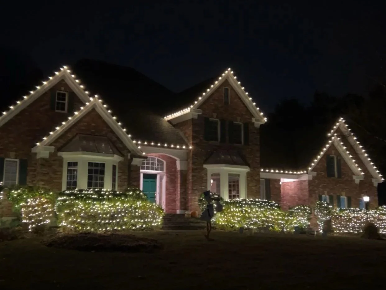 A large brick house decorated with white string lights along the rooflines and bushes in the yard at night.