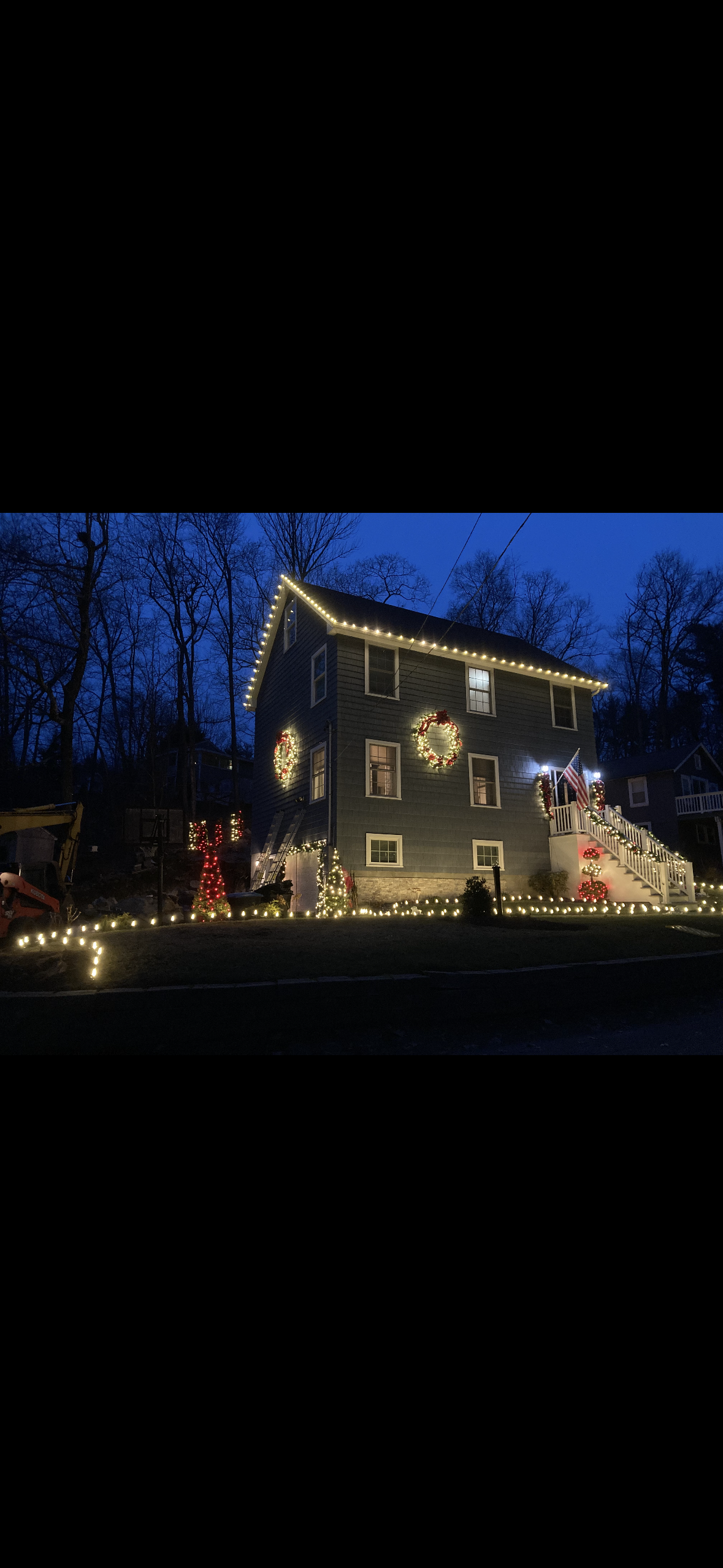 A house decorated with Christmas lights and wreaths, with a staircase adorned with lights and red poinsettias, during evening or early night.