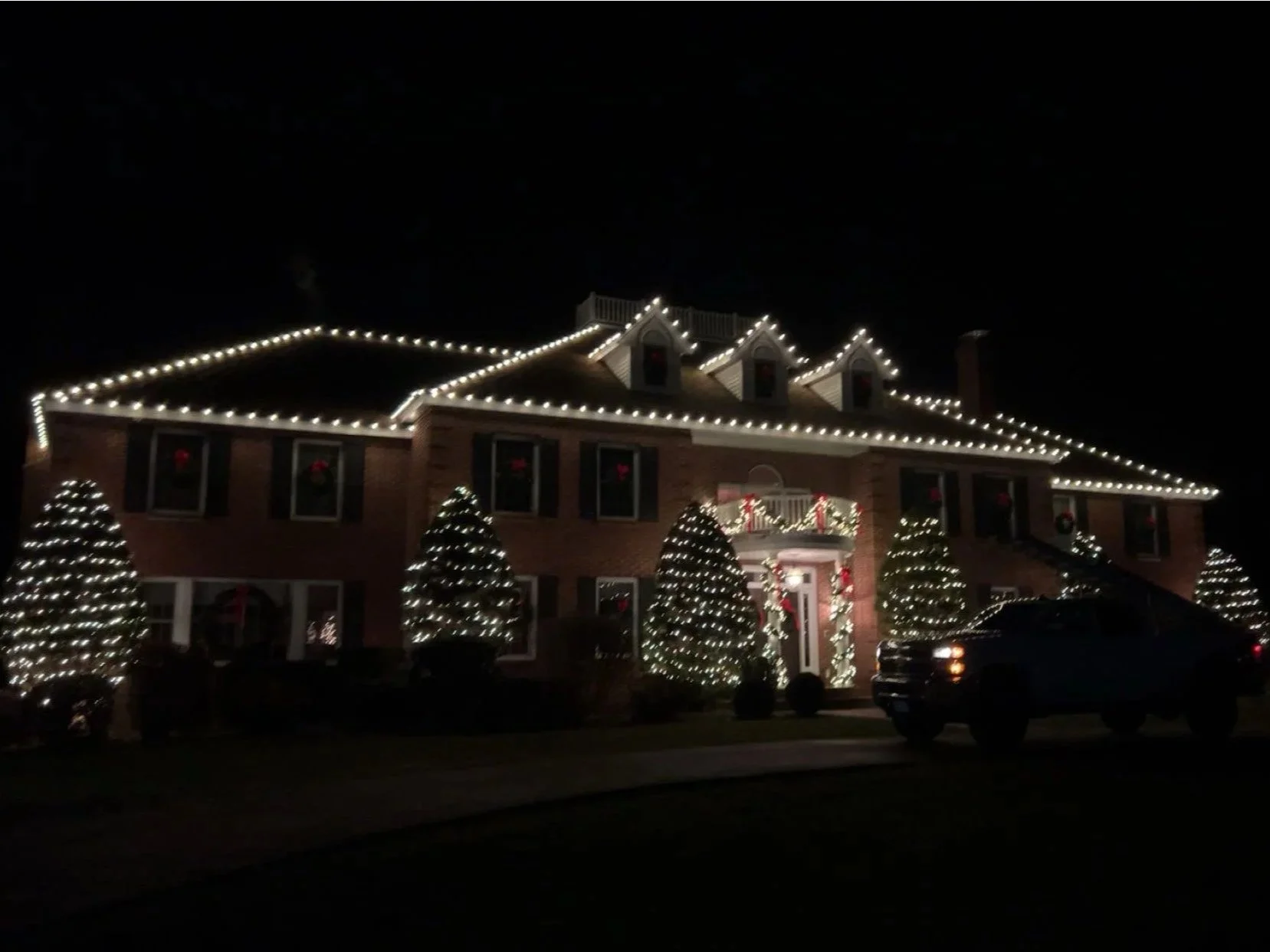 A large brick house decorated with Christmas lights outlining the roof and windows, with several Christmas trees decorated with string lights in the front yard, at night.