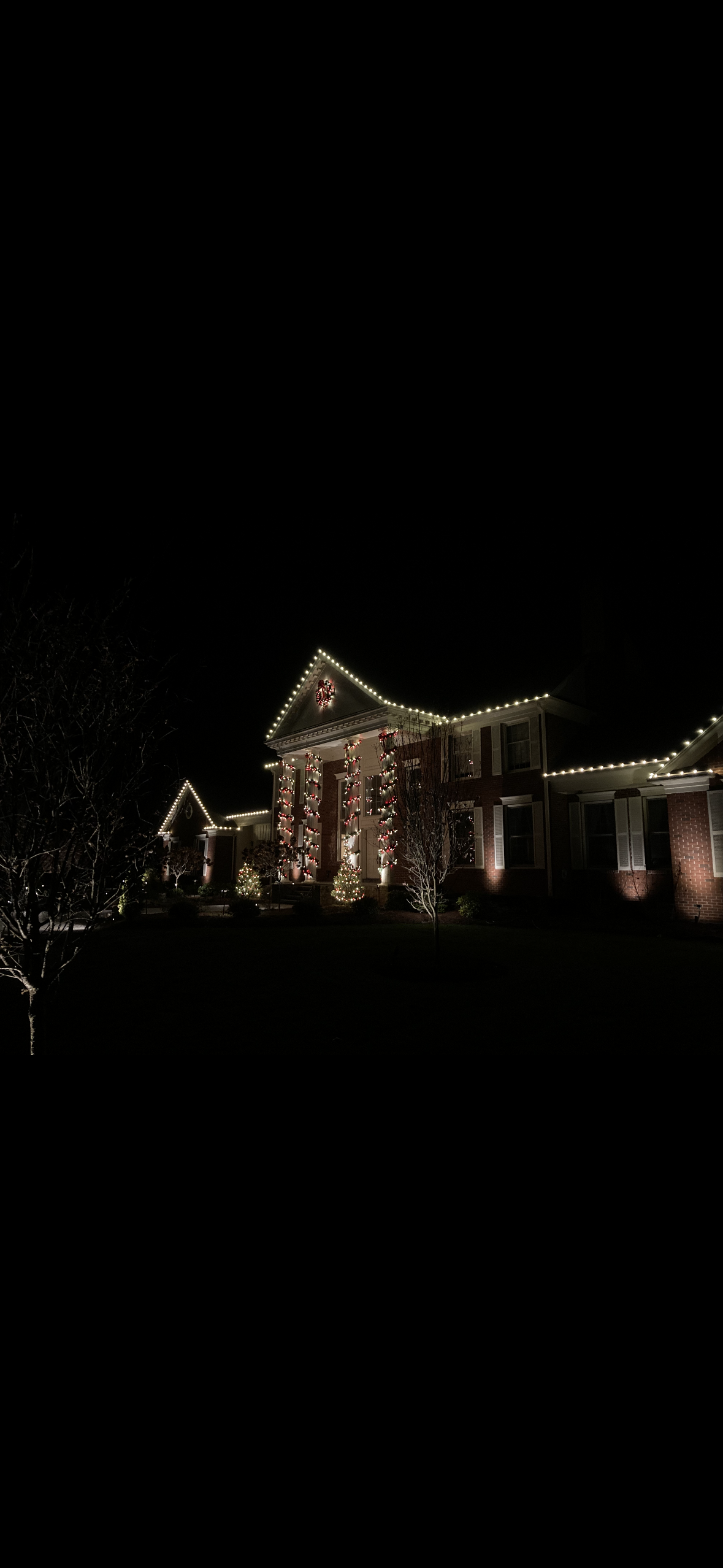 A house decorated with Christmas lights and ornaments at night, with a wreath in the gable and lit trees in the yard.