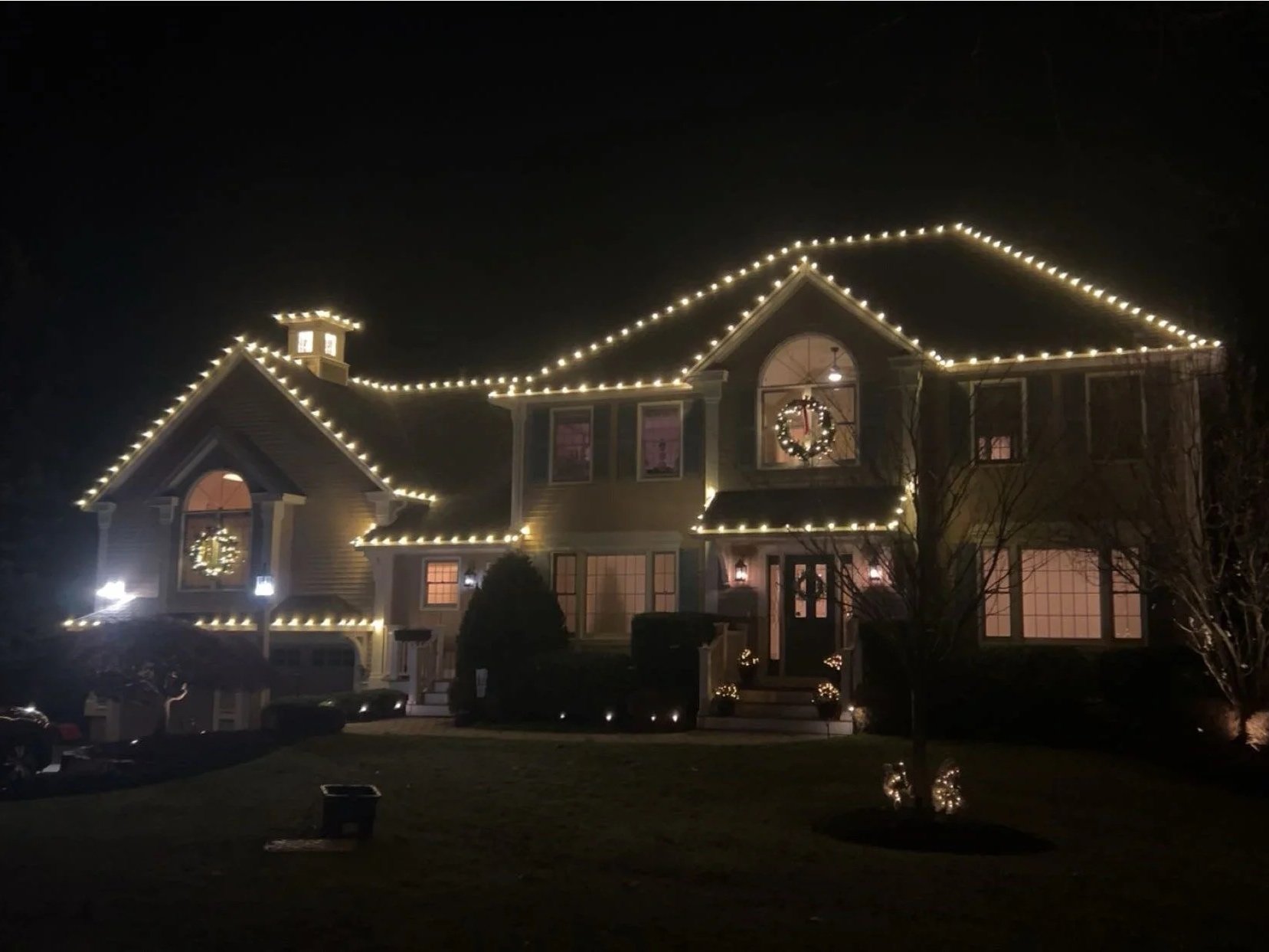 A house decorated with white Christmas lights outlining the roof and windows, with a festive wreath on the front door and illuminated decorations in the yard, during nighttime.