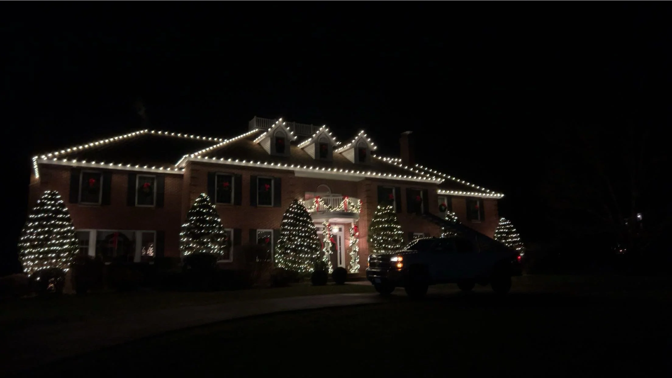 A large brick house decorated with bright white Christmas lights outlining the roof and windows, with illuminated Christmas trees and wreaths visible inside the windows and on the front porch, all illuminated against the night sky.