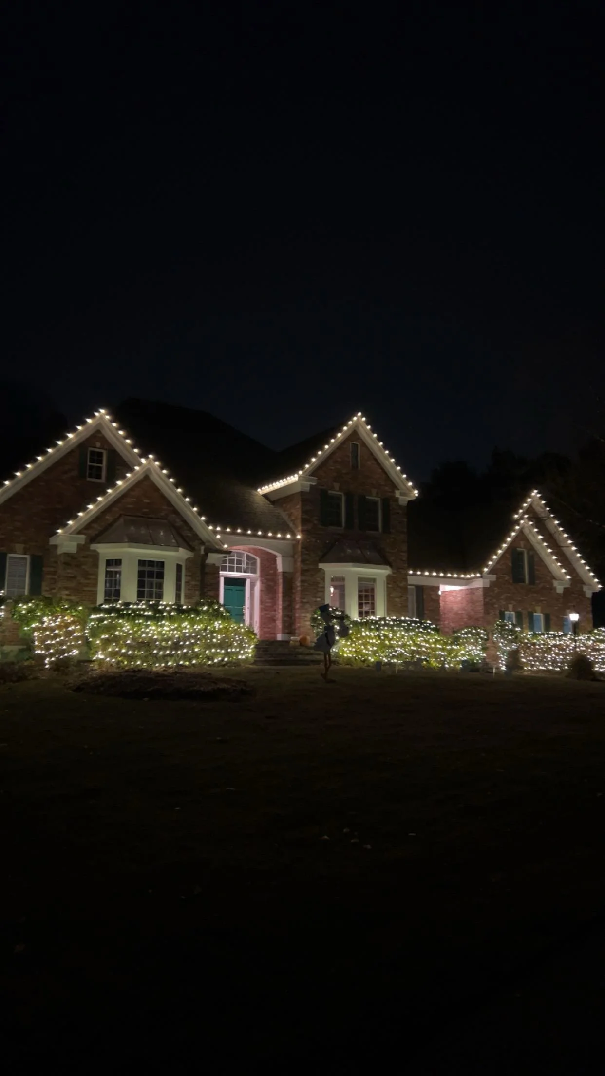 A large brick house decorated with white string lights along the rooflines and bushes in the yard at night.