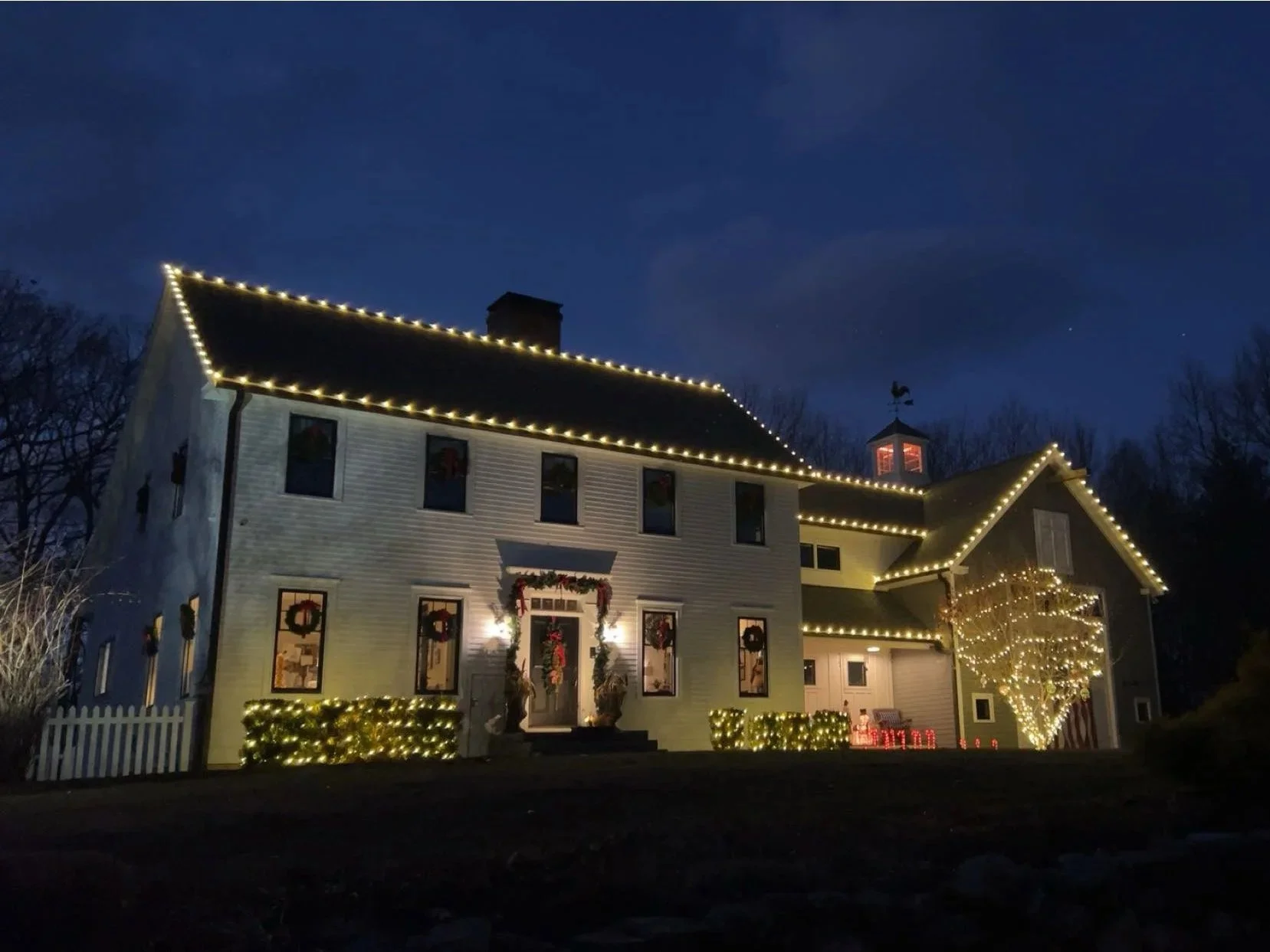A two-story house decorated with Christmas lights during nighttime. The house has string lights outlining the roof and a leafless tree wrapped in lights in front. The windows are decorated with wreaths and the front yard has illuminated bushes.