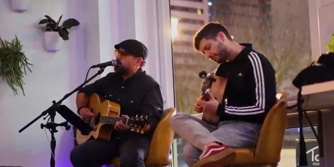 Two men are sitting and playing acoustic guitars in a room with large windows and indoor plants, with evening city lights visible outside.