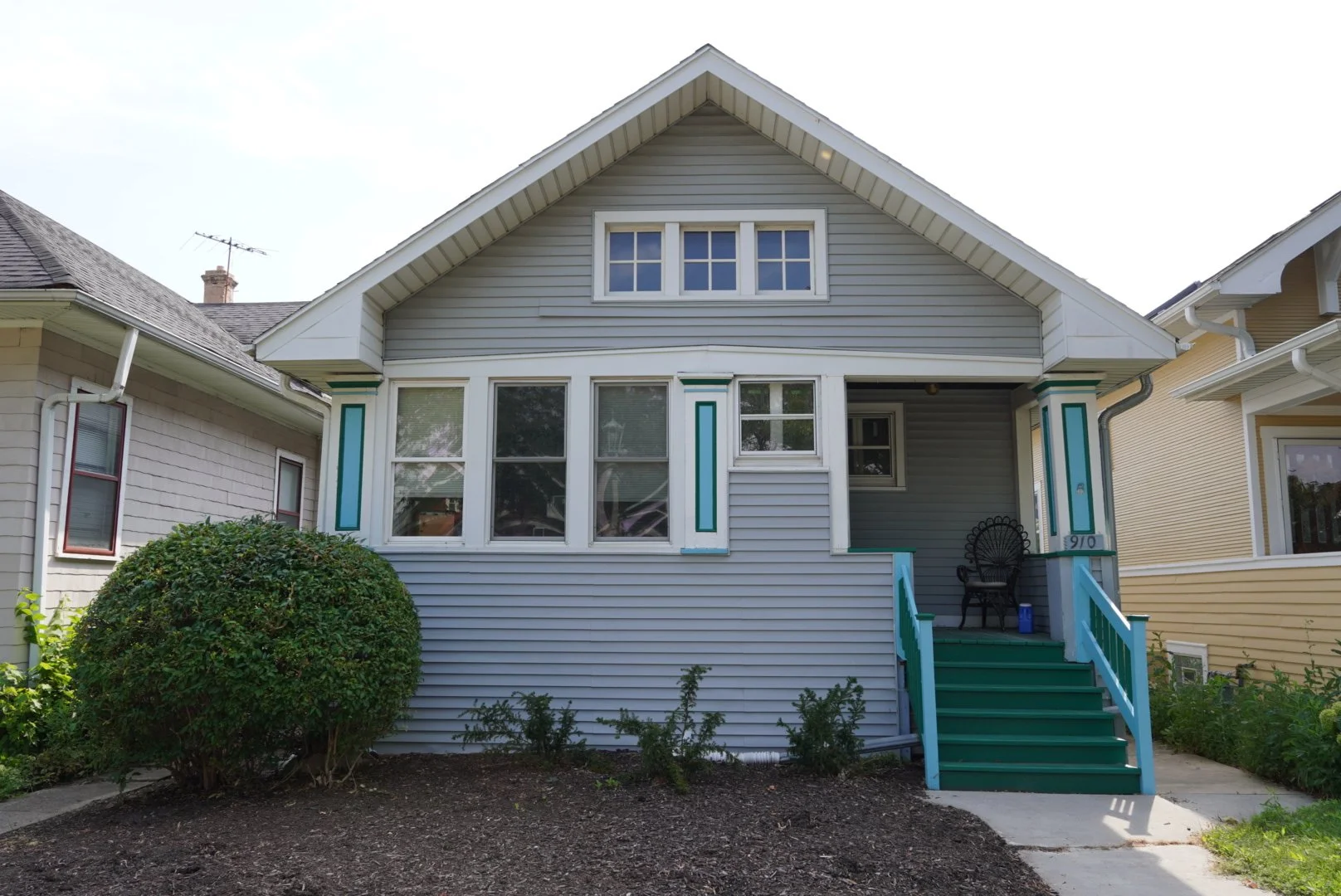 Front view of a light blue, two-story house with a gable roof, front stairs with teal railings, and a small porch with a black chair. There are neighboring houses and a shrub in front.