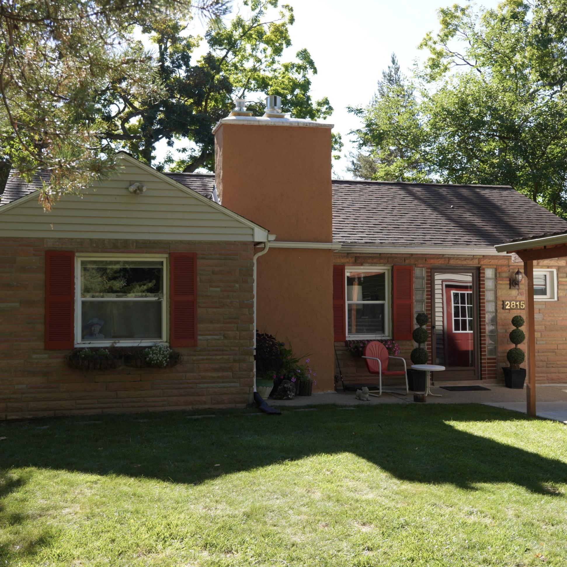 The back yard and patio of a house with a brick exterior, flowers, potted plants, a small table with a pink chair, and a large grassy lawn with trees in the background.