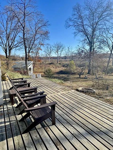 Wooden deck with four black rocking chairs overlooking a yard with leafless trees on a sunny day.