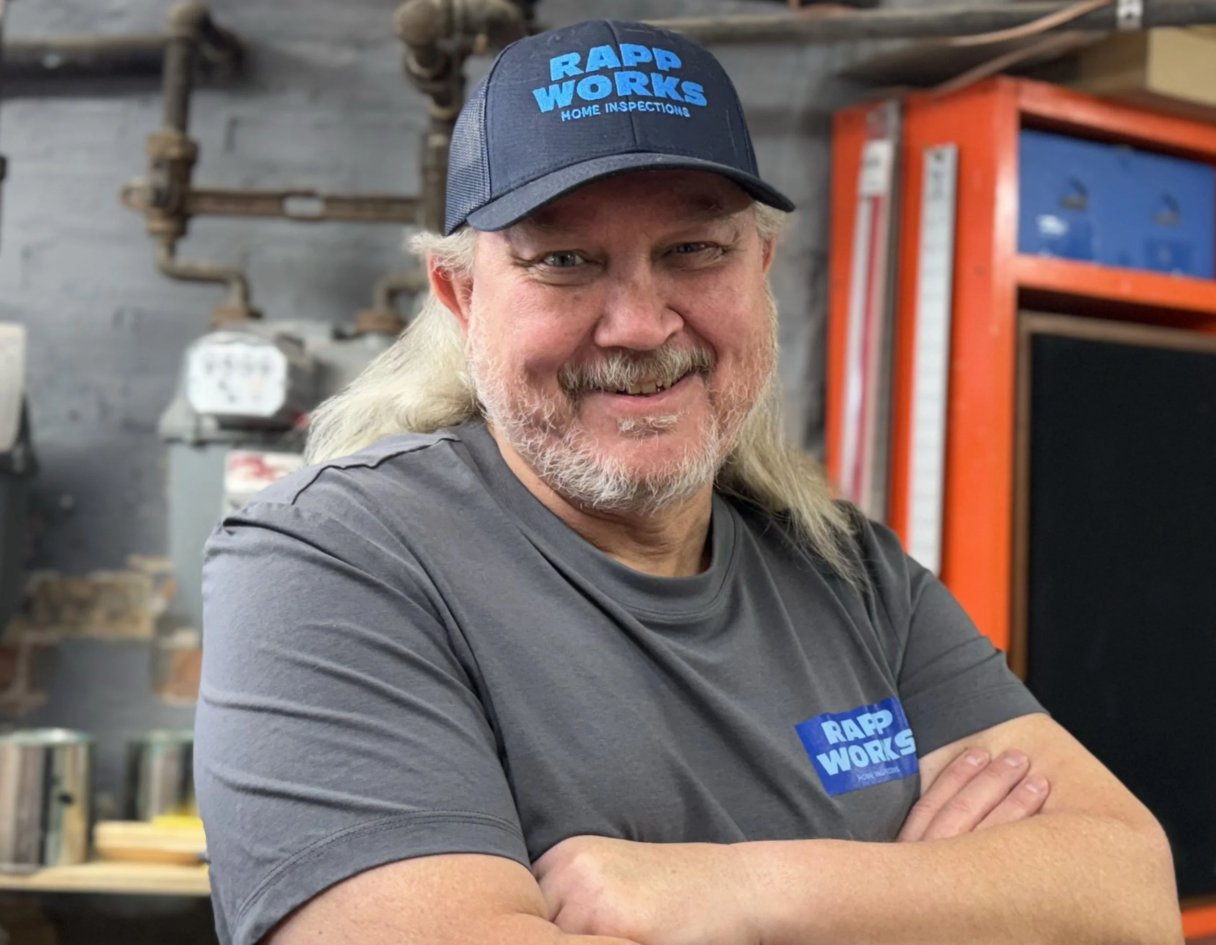 A man with long blond hair and a beard wearing a blue cap with the words 'RAPP WORKS HOME INSPECTIONS' in blue, smiling, standing inside a workshop with tools and equipment in the background.