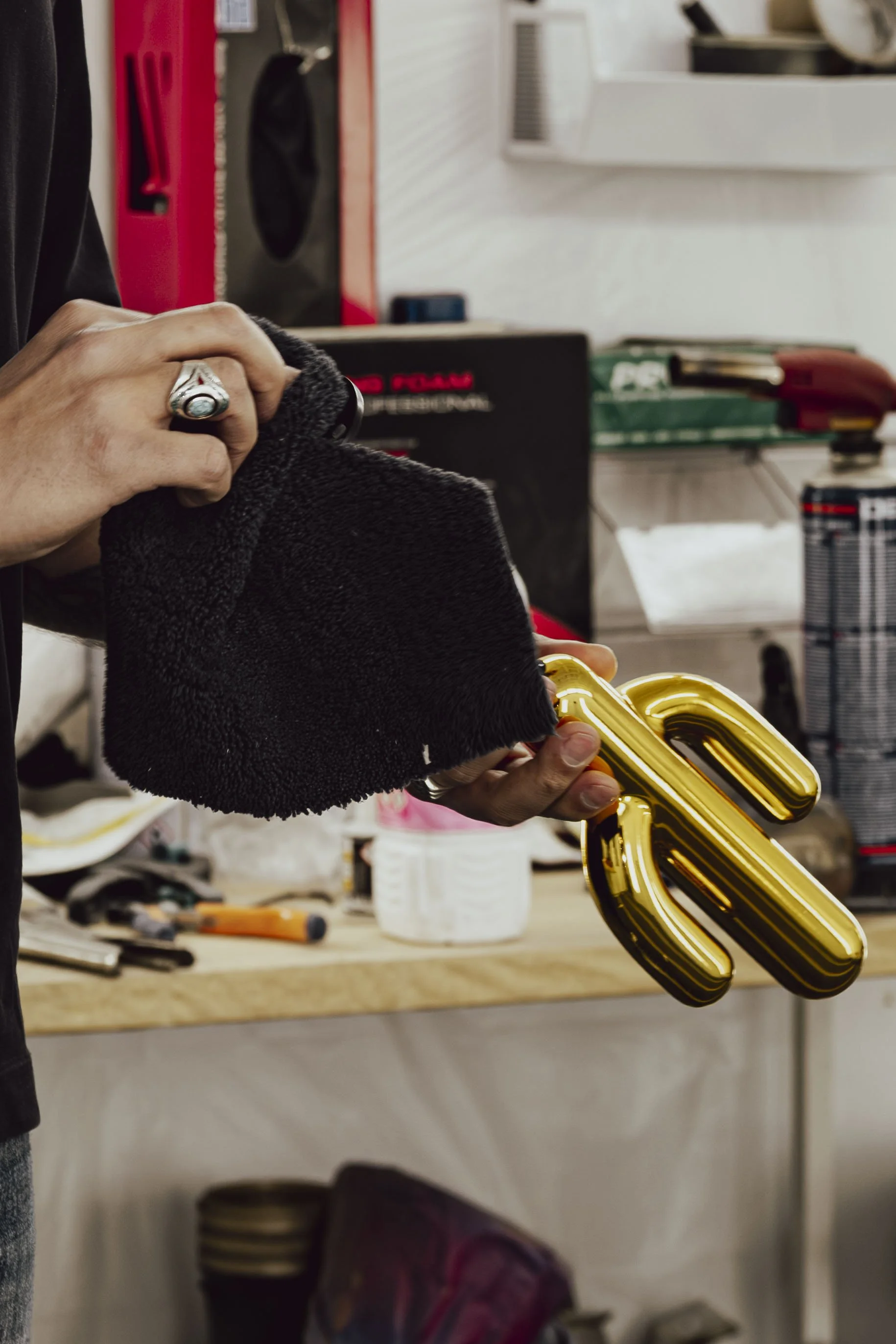 Person holding a gold metallic number 2 sign in one hand and a black polishing cloth in the other, in a workshop setting.