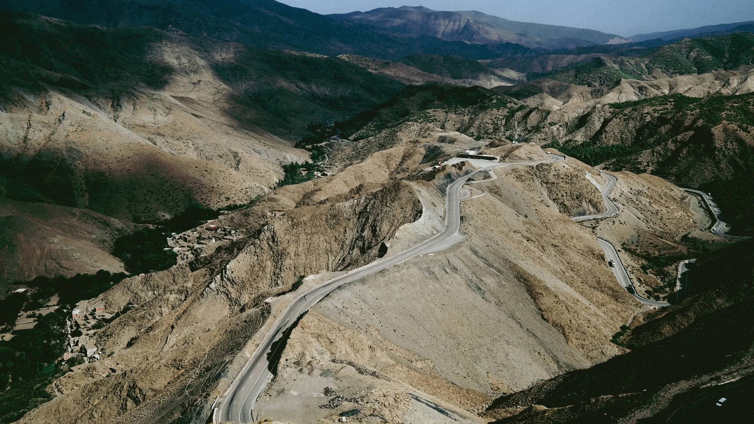 A winding mountain road cutting through dry, rugged terrain in a mountainous landscape with distant mountain ranges in the background.