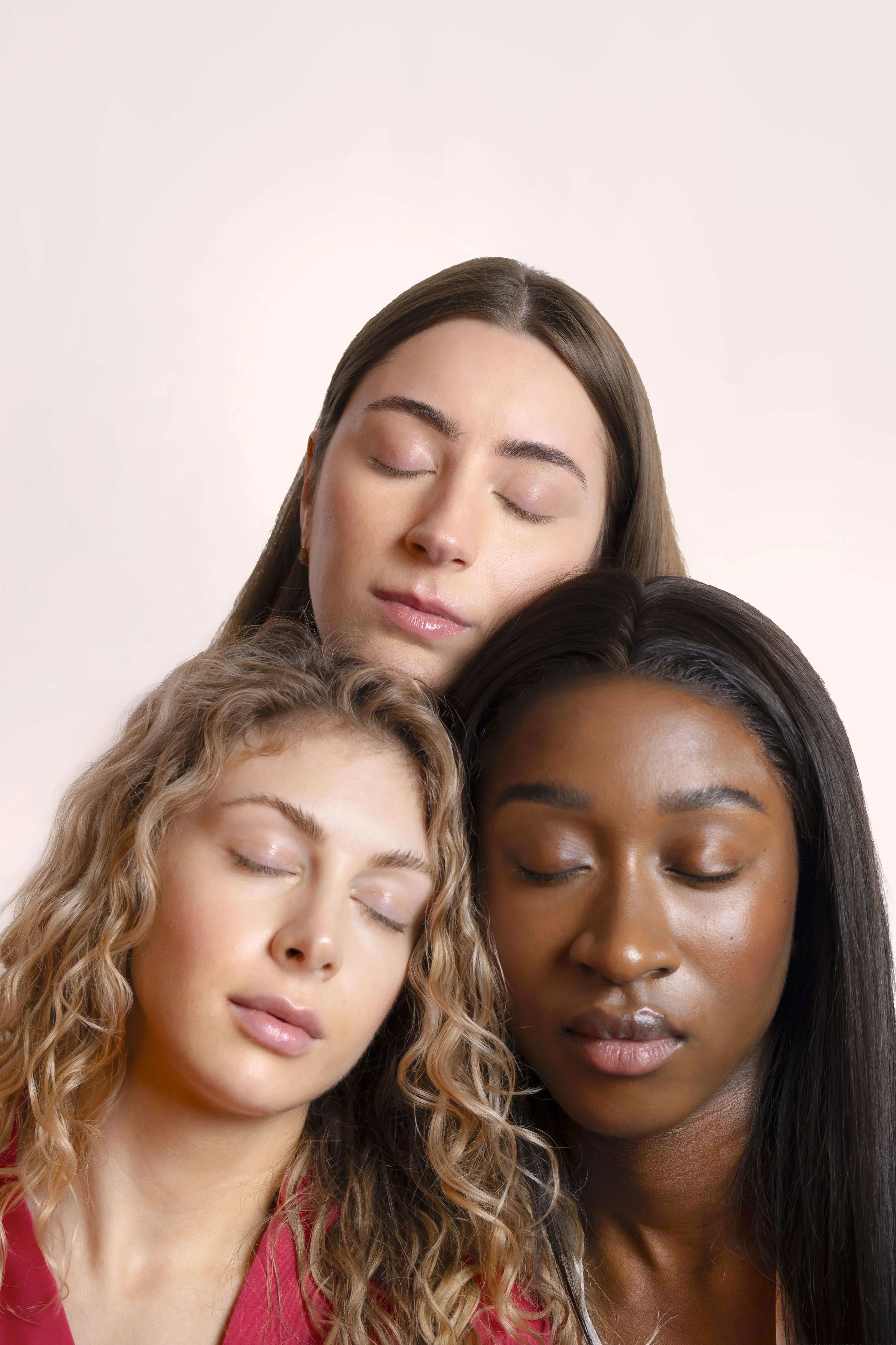 Three women with different skin tones and hair colors have their eyes closed and are touching foreheads and heads in a group hug.