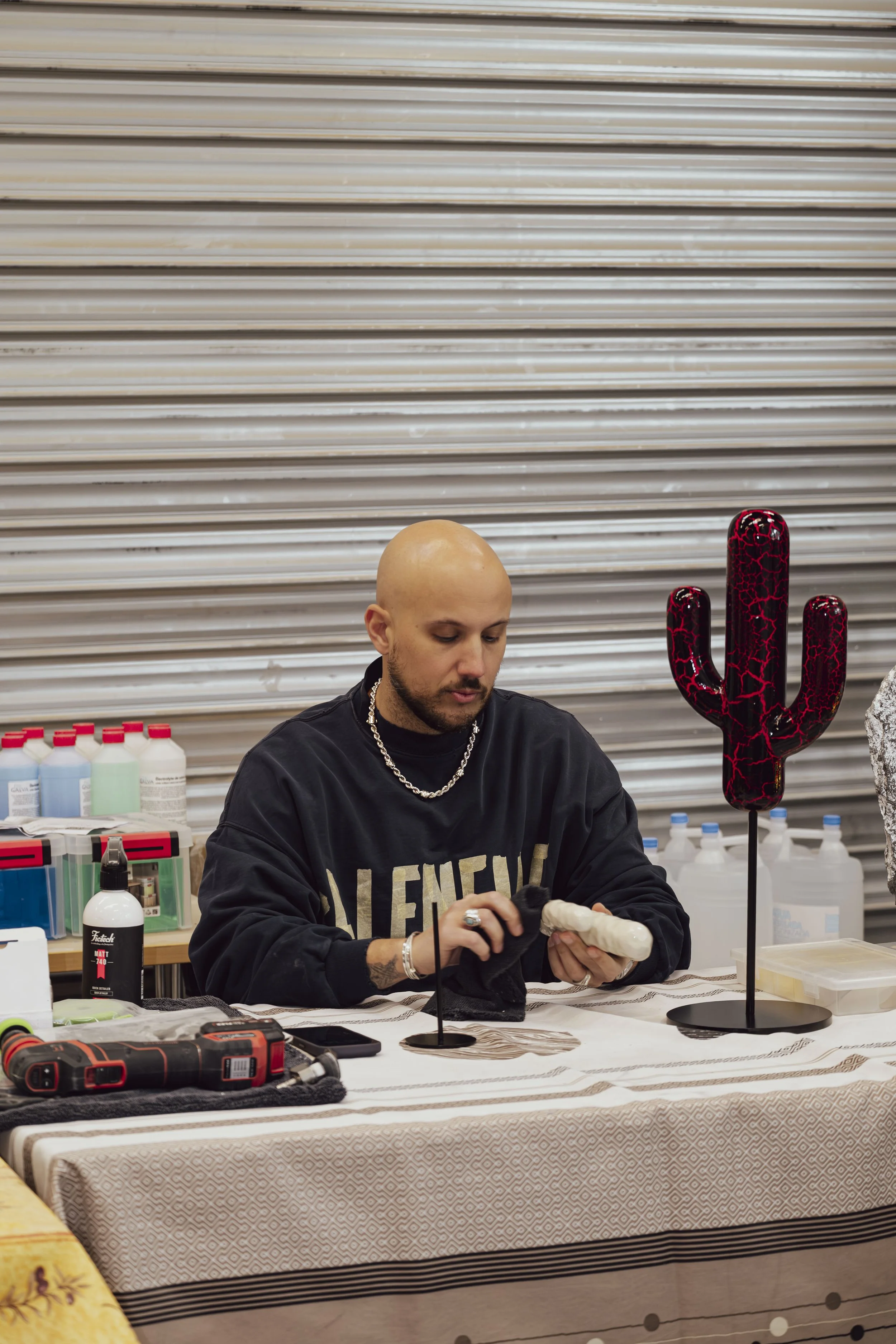 A man sits at a table, working with crafting tools surrounded by various supplies. There are two cactus-shaped sculptures on stands, one of which is black and red with a cracked pattern.
