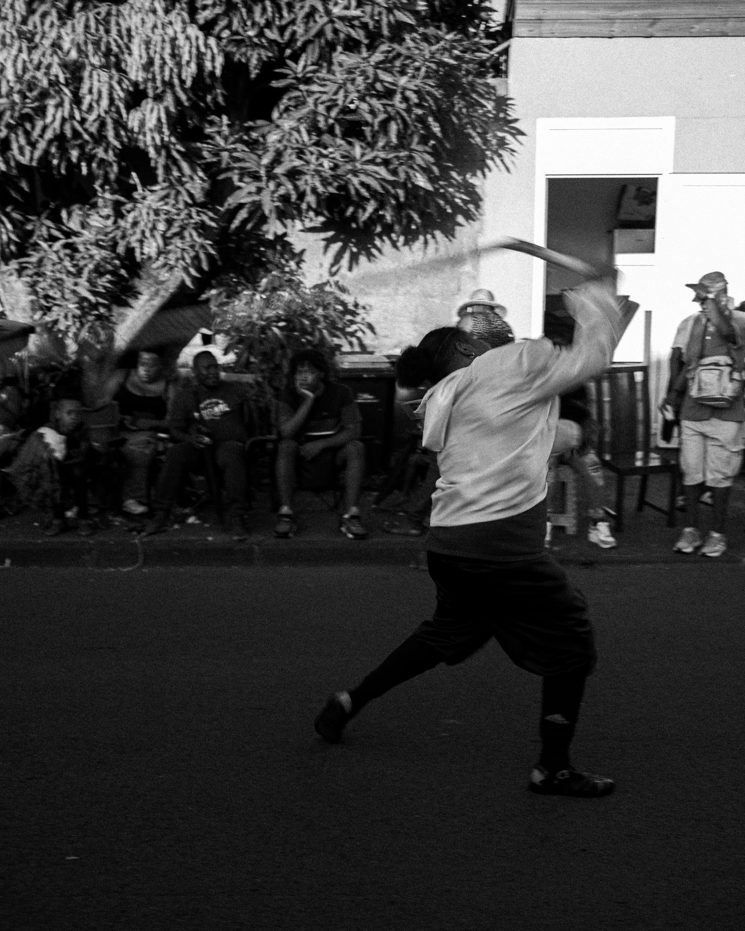 A person swinging a stick or bat in an outdoor setting with a group of people watching, some seated and some standing, in the background.