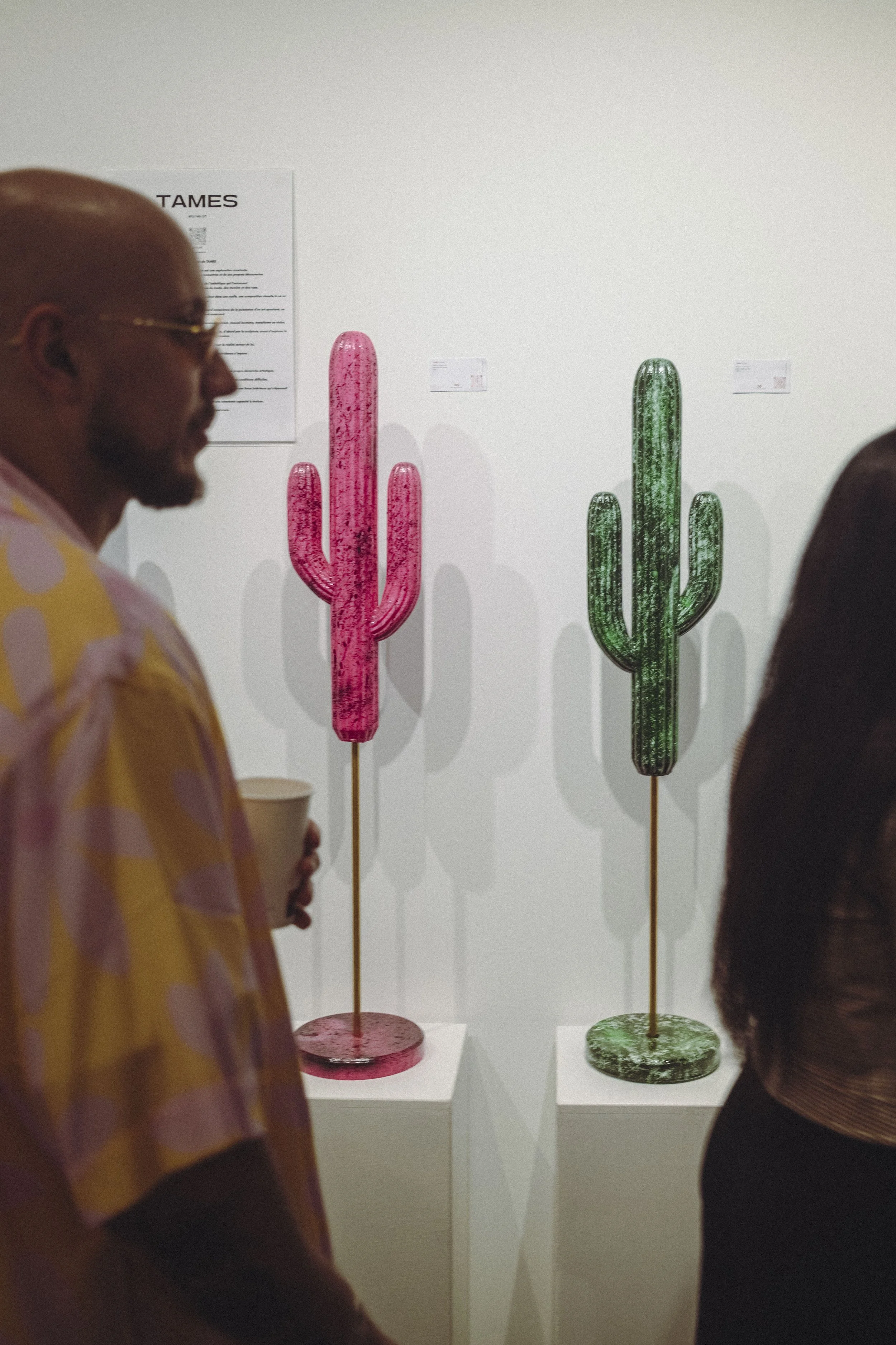 Two cactus-shaped sculptures, one pink and one green, displayed on white pedestals at an art gallery.