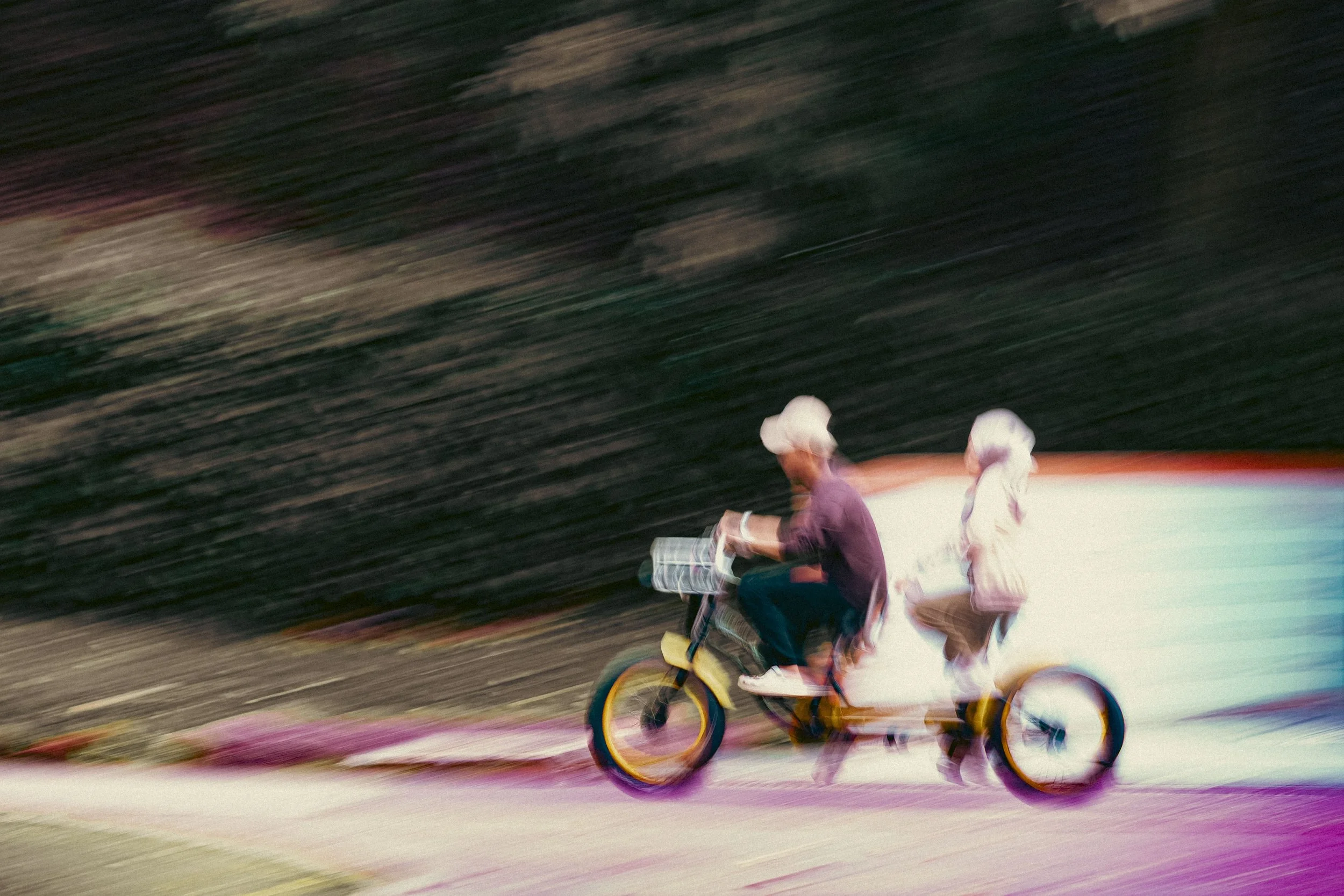 A person riding a tricycle with a child seated behind them, moving quickly along a colorful street.