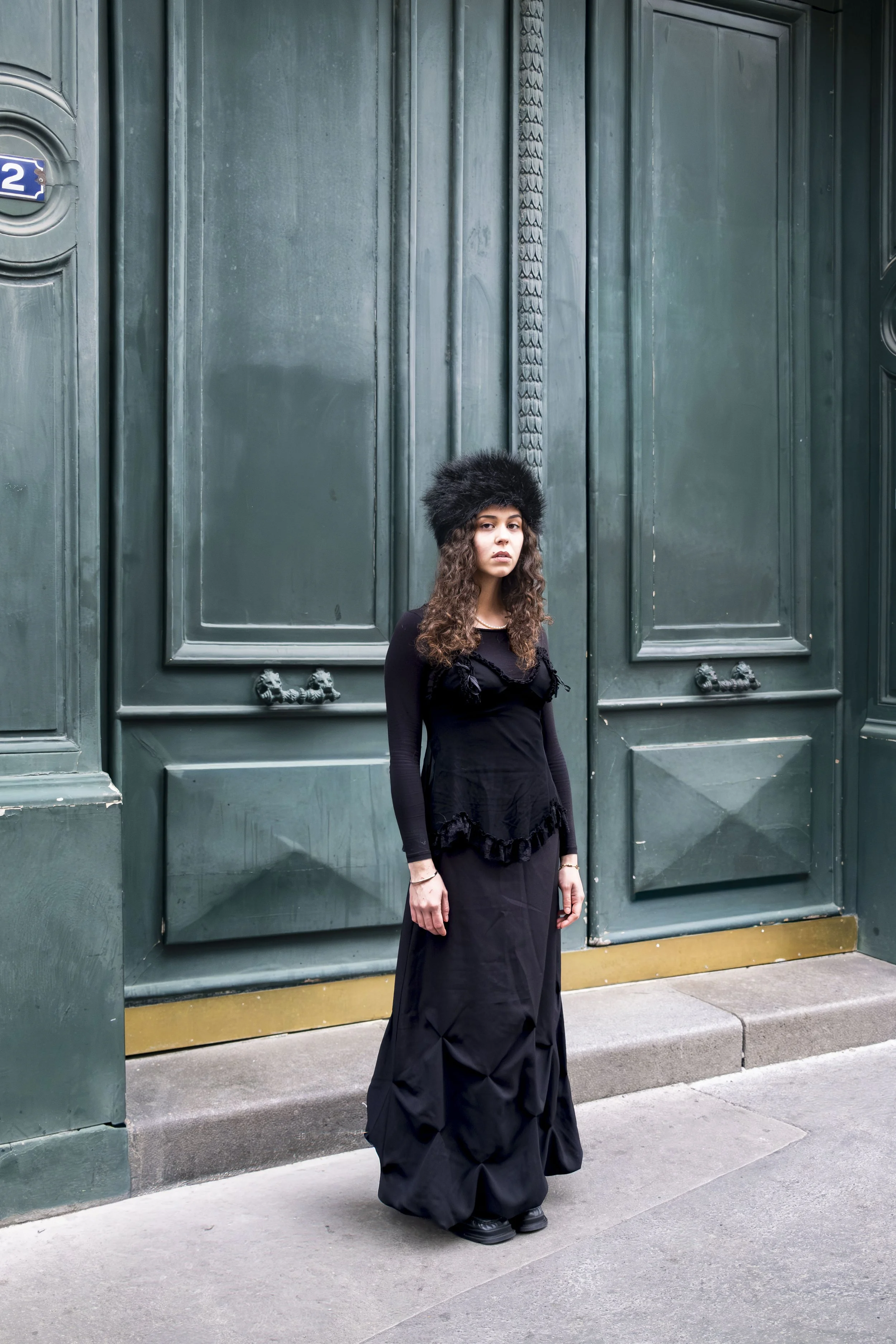 Woman standing in front of a large, dark green door with ornate detailing, dressed in black with a furry hat and long skirt.