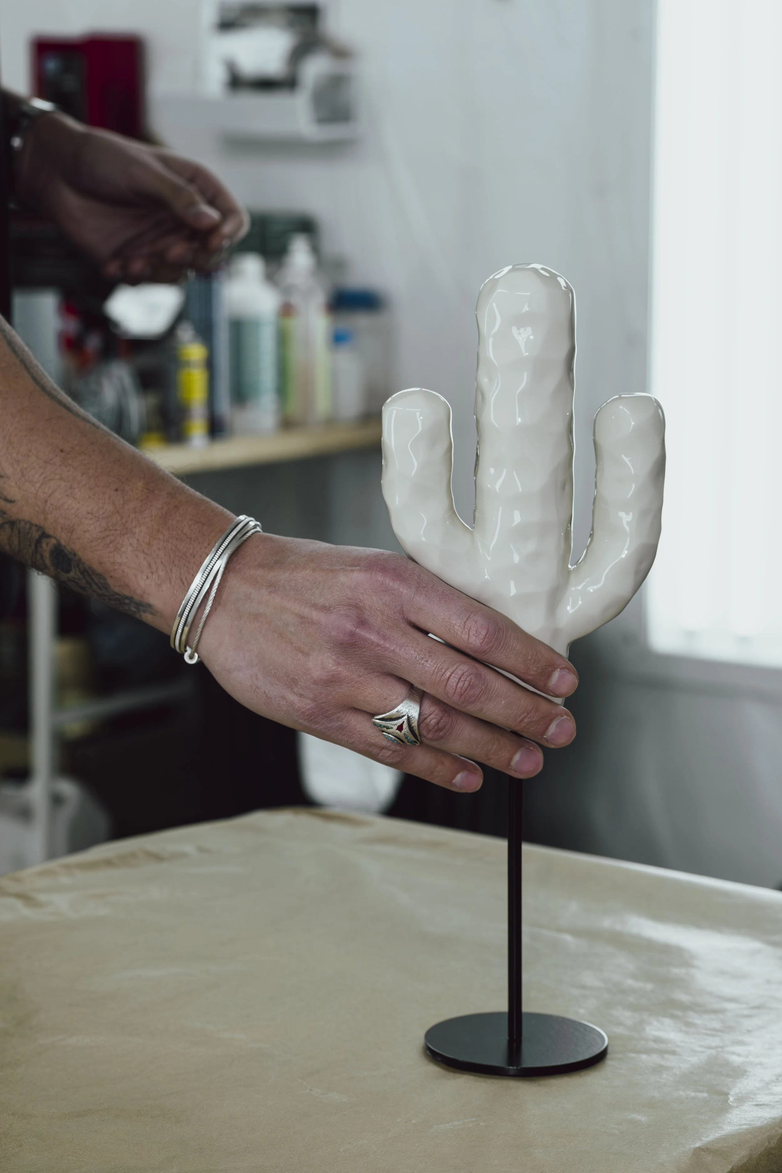 Hands holding a white ceramic cactus sculpture on a black stand, with a background of art supplies and a window.