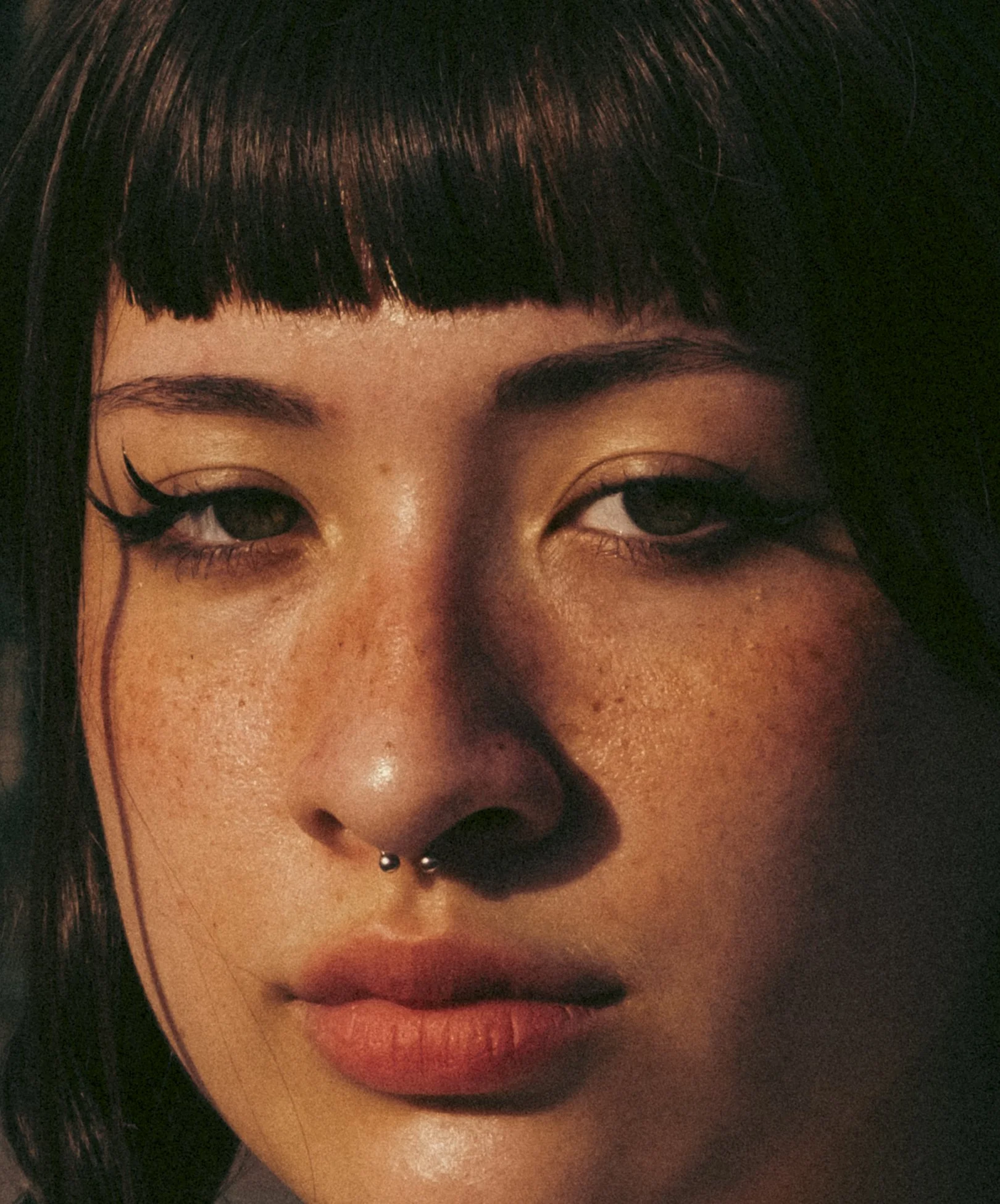 Close-up of a woman with dark hair, freckles, and a septum piercing, wearing makeup with winged eyeliner, in dim lighting.