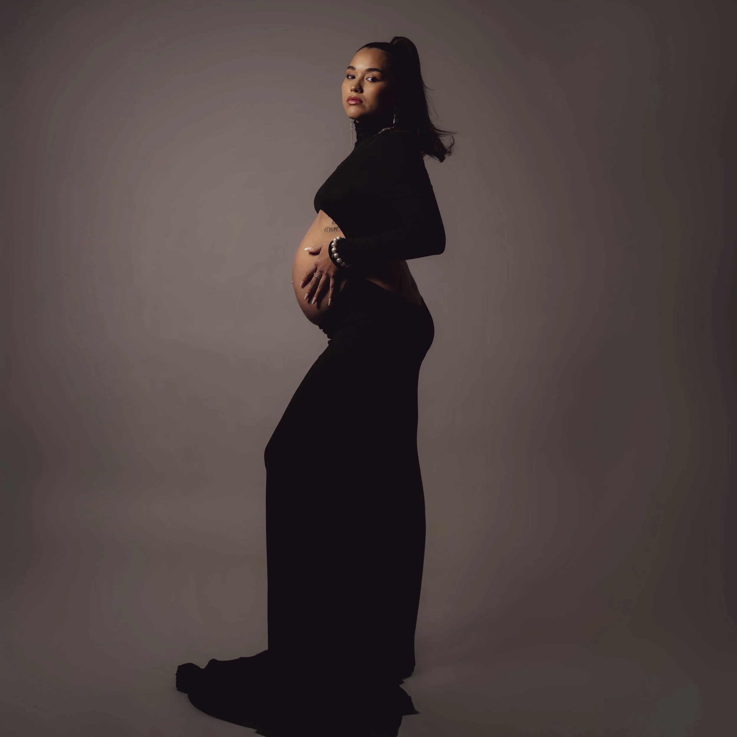 A pregnant woman with dark hair in a high ponytail, wearing black clothing, standing sideways against a plain neutral background, with her hand on her belly.